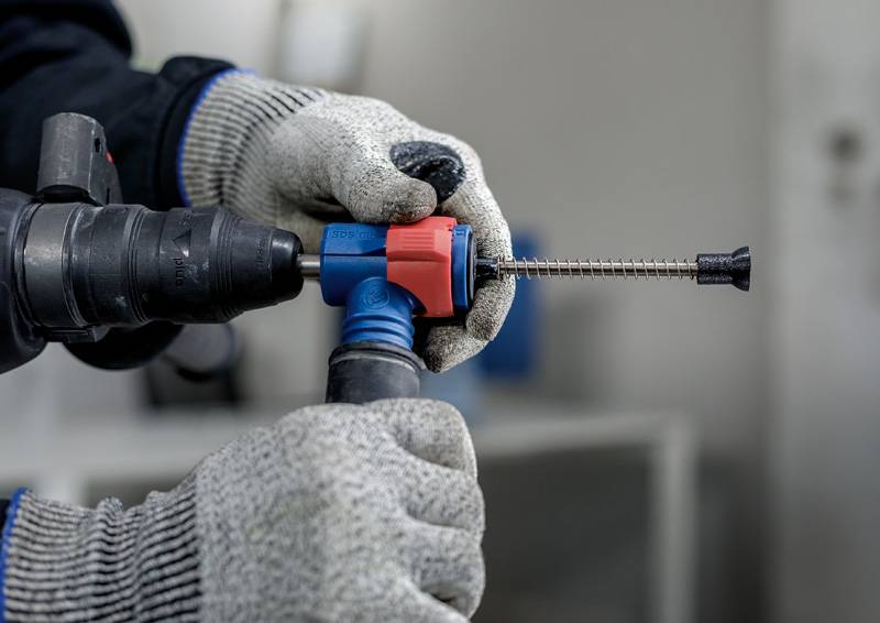 Nahaufnahme von Händen in Arbeitshandschuhen, die eine Bohrmaschine bedienen. Ein Einsatzstück wird sicher festgehalten.