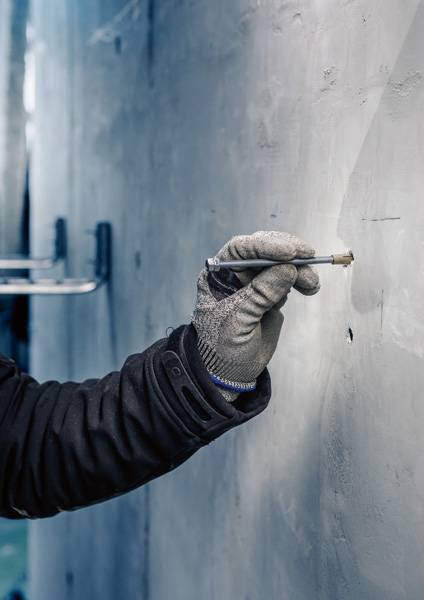 Eine behandschuhte Hand hält ein Stiftwerkzeug und zeichnet auf eine graue Wand, möglicherweise Teil eines industriellen Arbeitsprozesses.