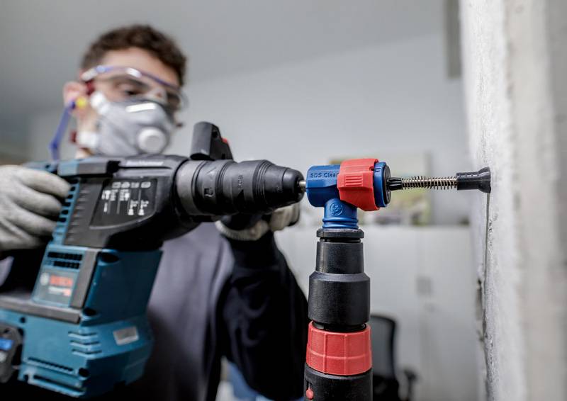 Person trägt Schutzbrille und Atemmaske, nutzt eine Bohrmaschine, um in eine Betonwand zu bohren. Sicherheit steht im Vordergrund.