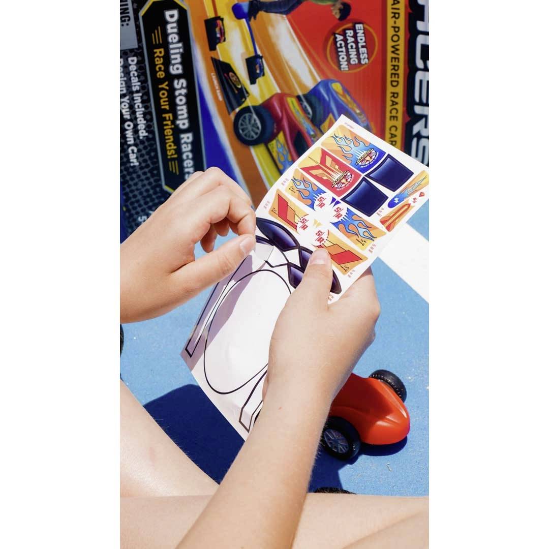 Ein Kind hält Aufkleber und ein kleines Spielzeugauto mit einer orangefarbenen Karosserie, während eine Verpackung eines Spielzeugautos im Hintergrund liegt.