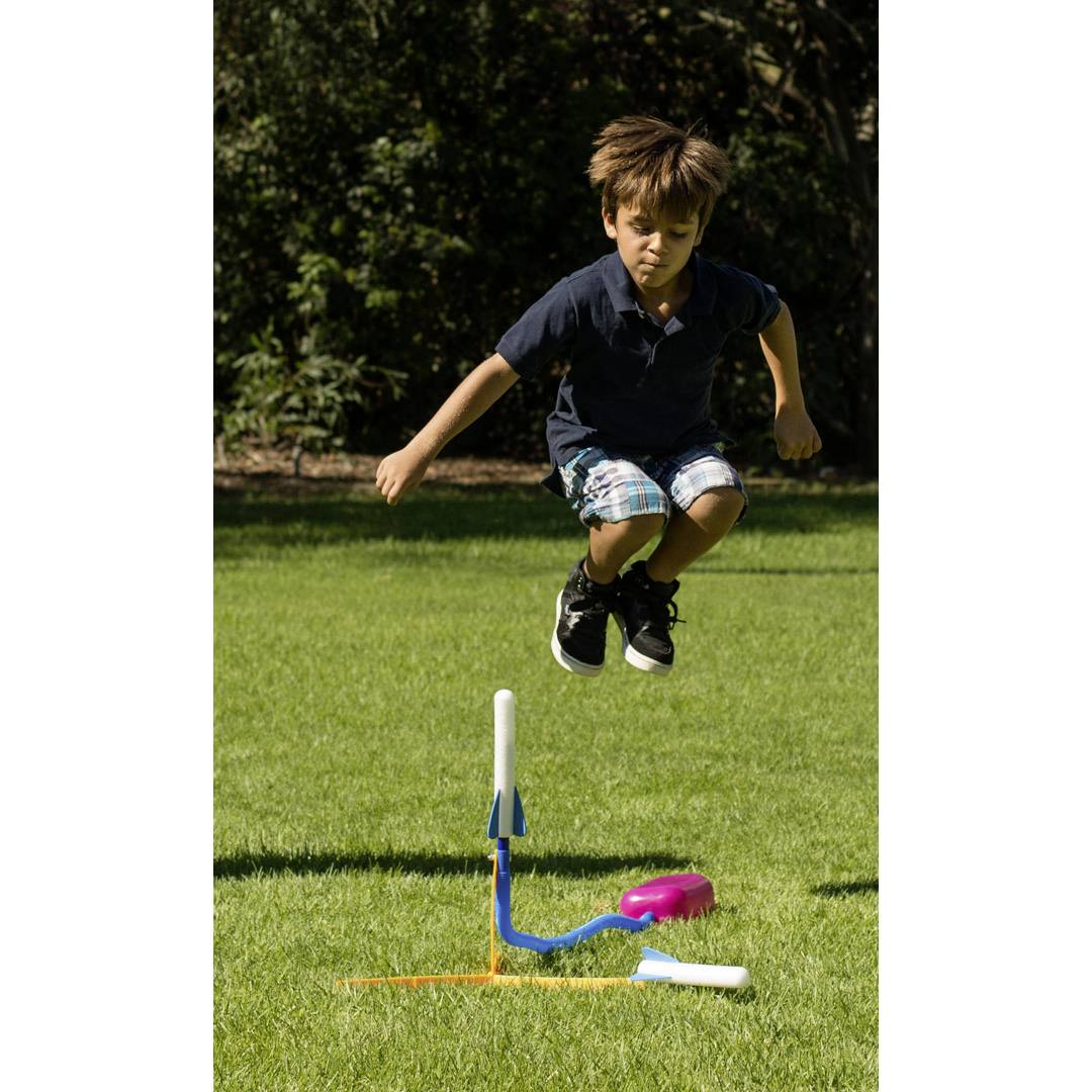 Ein Junge springt kraftvoll in die Luft und landet auf einer Tretpumpe, die eine Rakete aus Plastik vertikal in die Luft schießt.