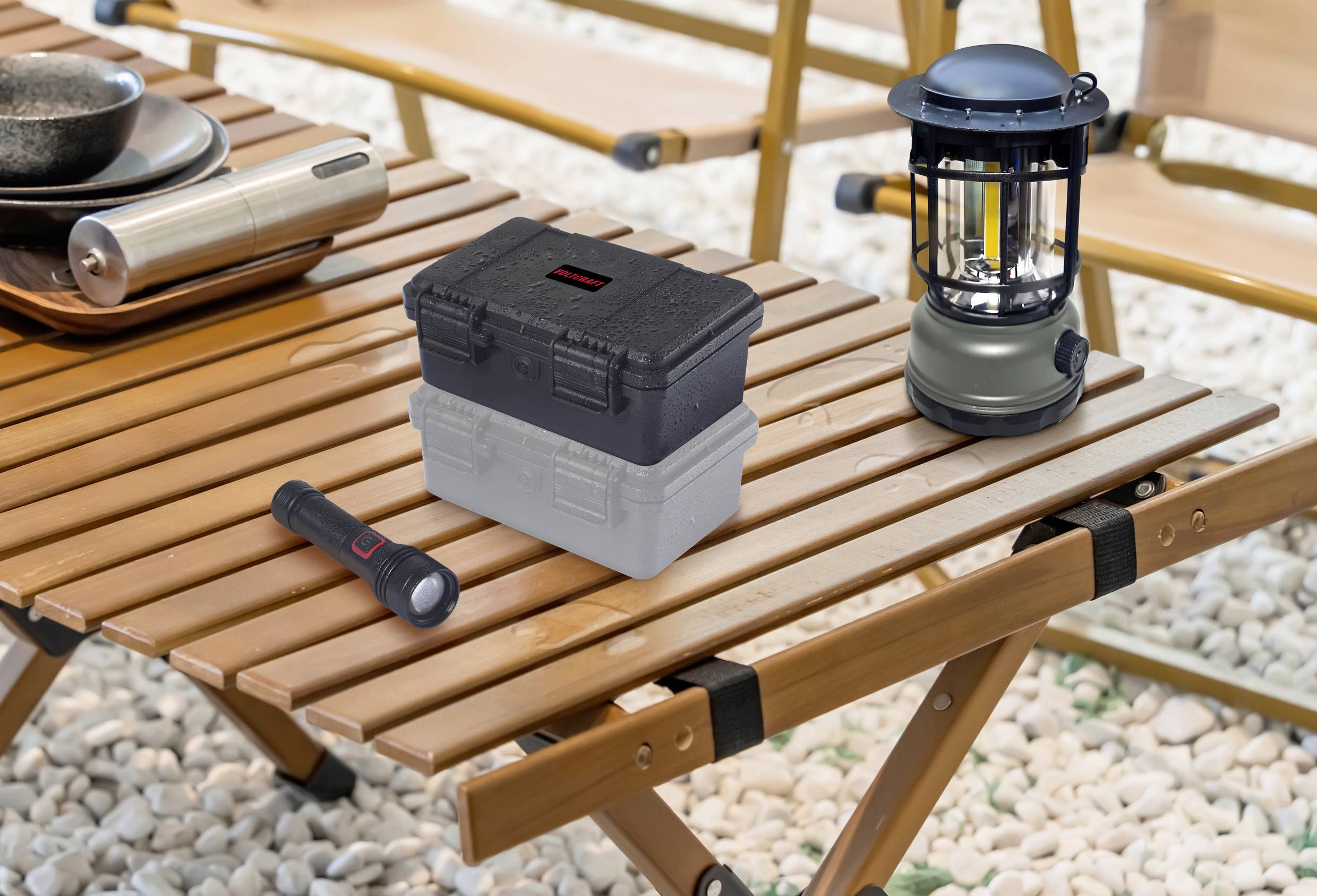Ein klappbarer Holztisch im Freien mit einer Laterne, einer Taschenlampe, einem Alubehälter und einer Kiste darauf, umgeben von Kiesboden.