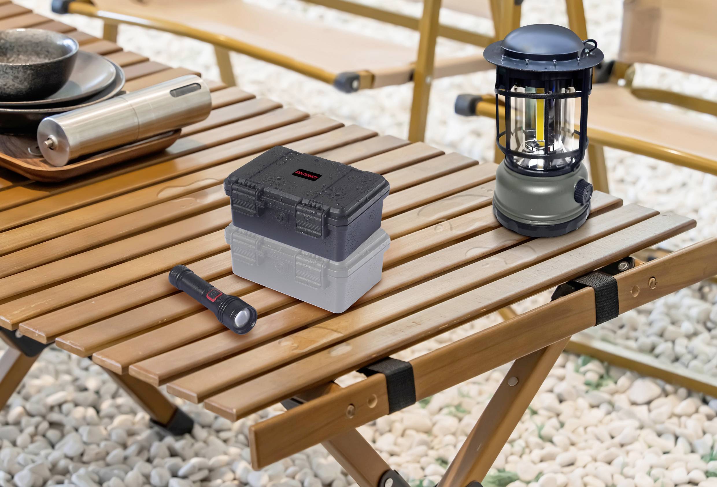 Campingtisch aus Holz mit einer Laterne, einer Taschenlampe und einem kleinen, stapelbaren Behälter. Im Hintergrund sind Klappstühle.
