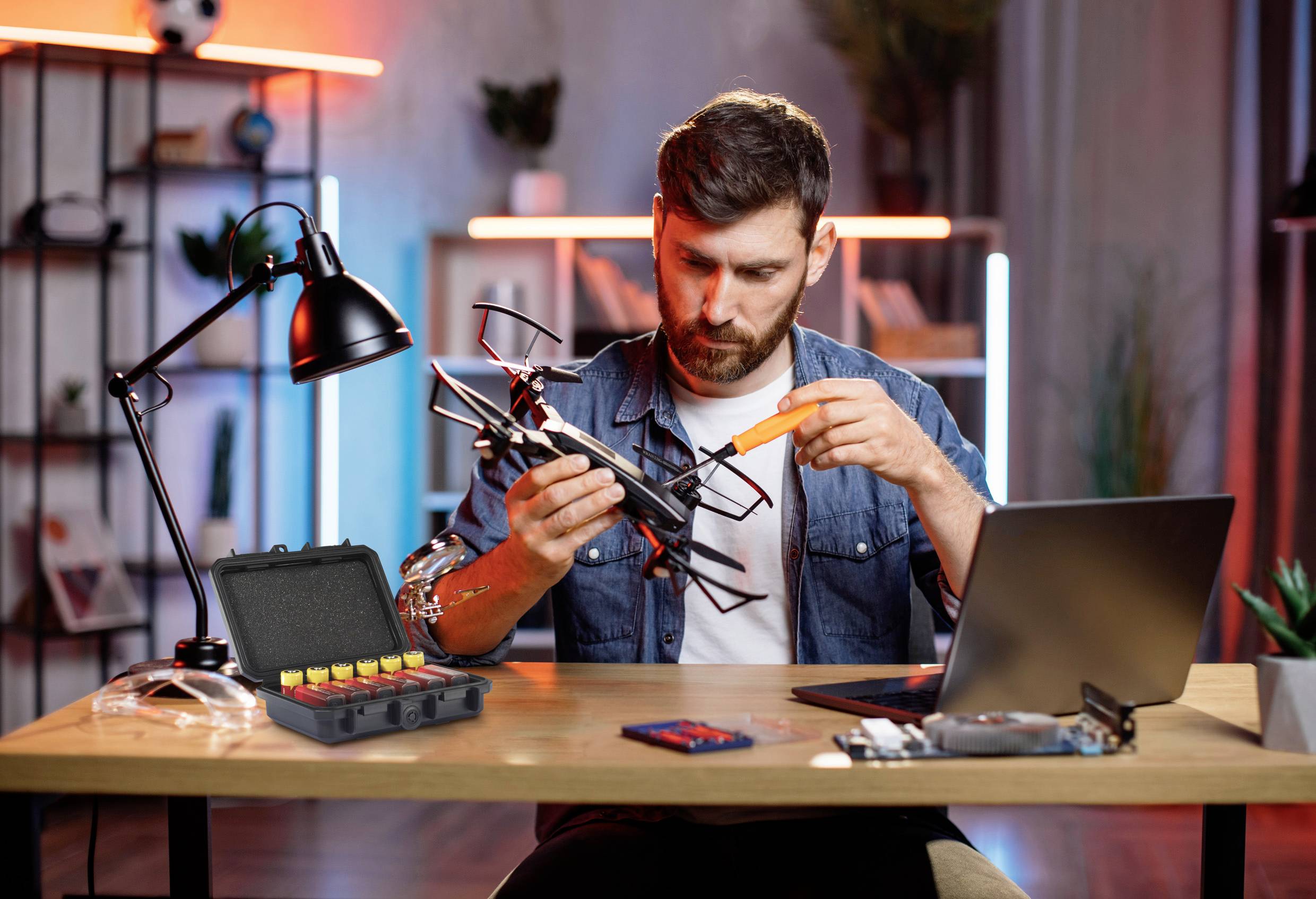 Ein Mann sitzt an einem Tisch und repariert eine Drohne. Neben ihm ist ein Laptop und ein offener Werkzeugkoffer mit Batterien.