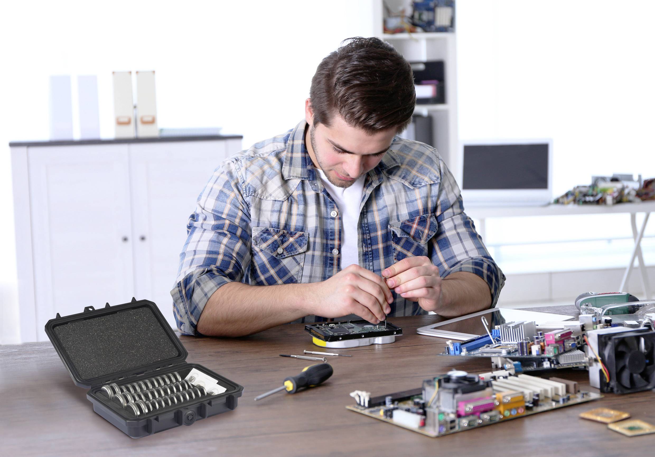 Ein Mann repariert ein elektronisches Gerät an einem Schreibtisch. Auf dem Tisch liegen Werkzeuge und elektronische Bauteile. Im Hintergrund sind Regale und Zubehör zu sehen.