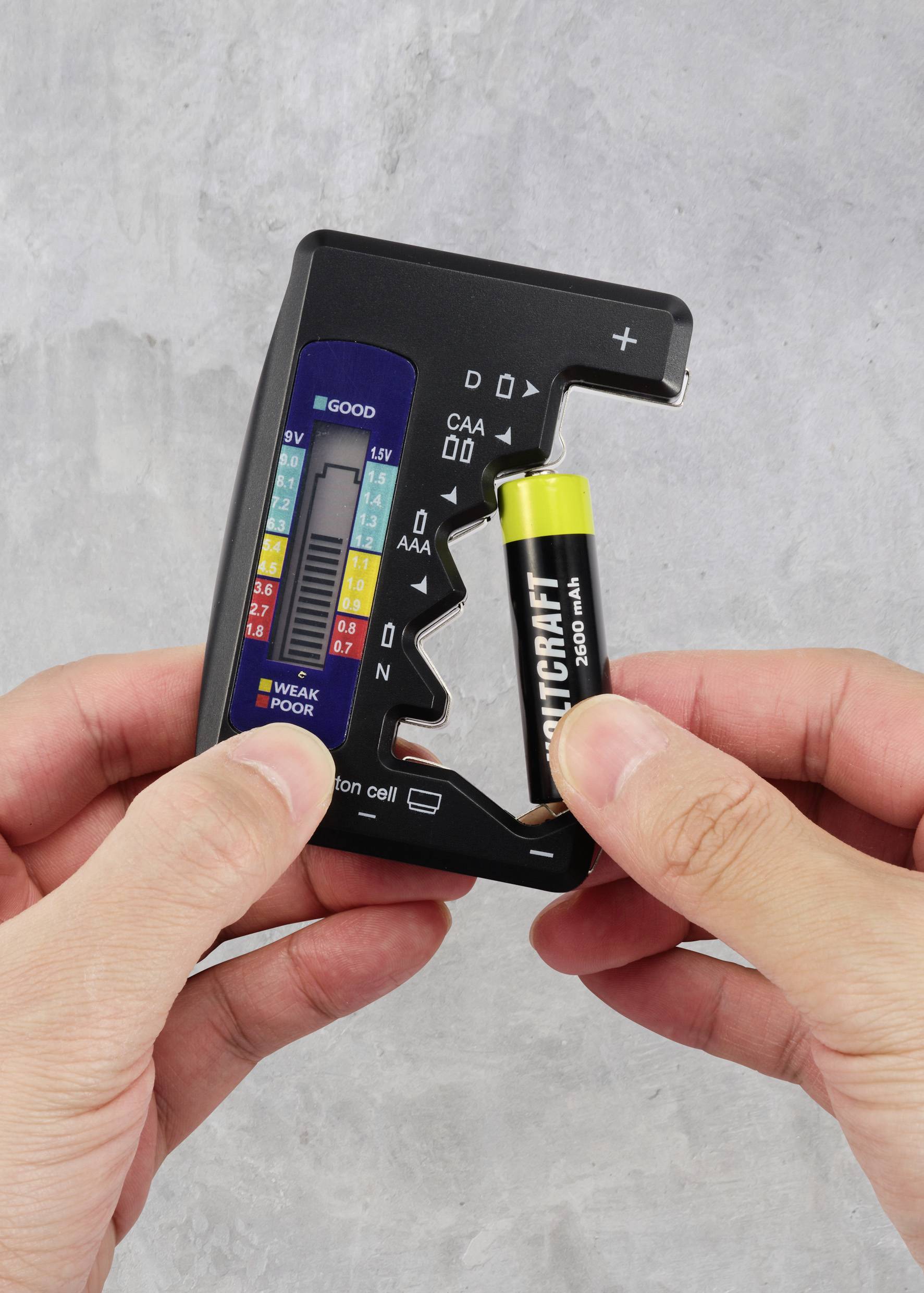 Eine Person hält einen Batterietester und testet eine AA-Batterie. Der Tester zeigt gute Leistung an. Hintergrund grau.