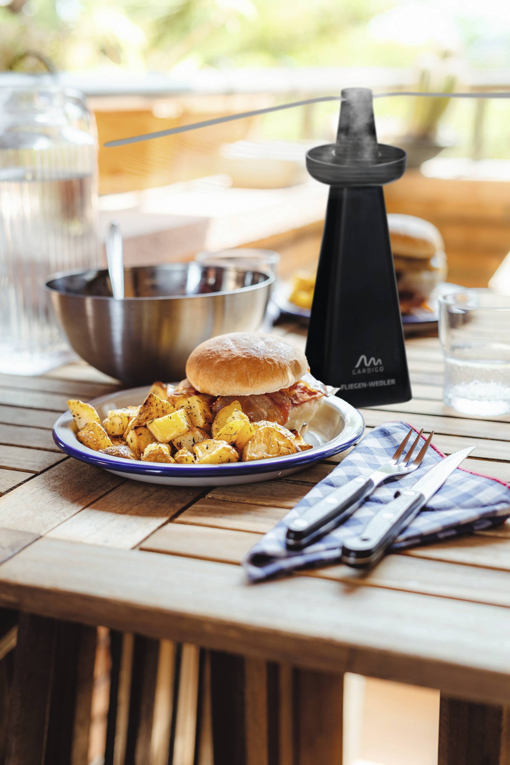 Ein Tisch auf einer Terrasse mit einem Teller Pommes und einem Burger, daneben ein Wasserkrug, eine Metallschüssel und ein Glas.
