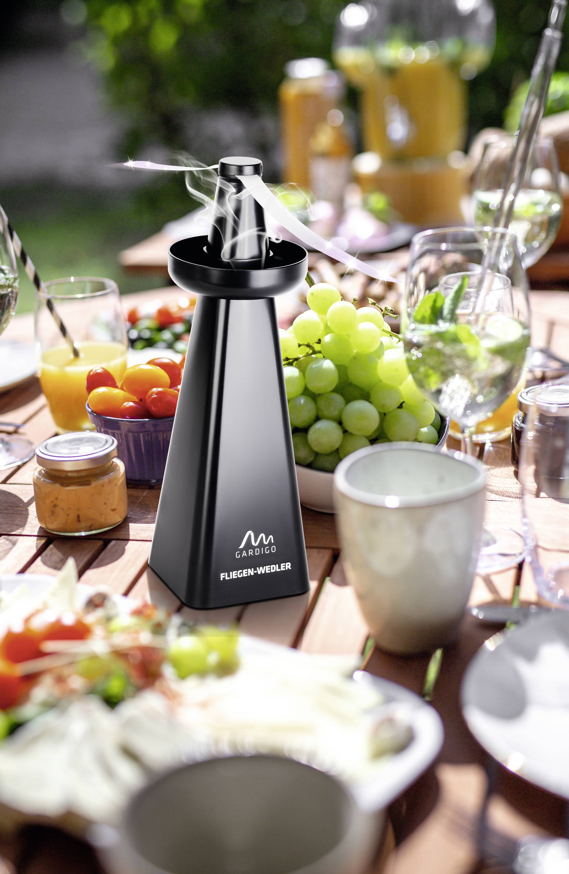 Schwarze elektronische Pfeffermühle auf gedecktem Gartentisch mit Speisen und Getränken. Im Hintergrund frisches Obst und Saft.