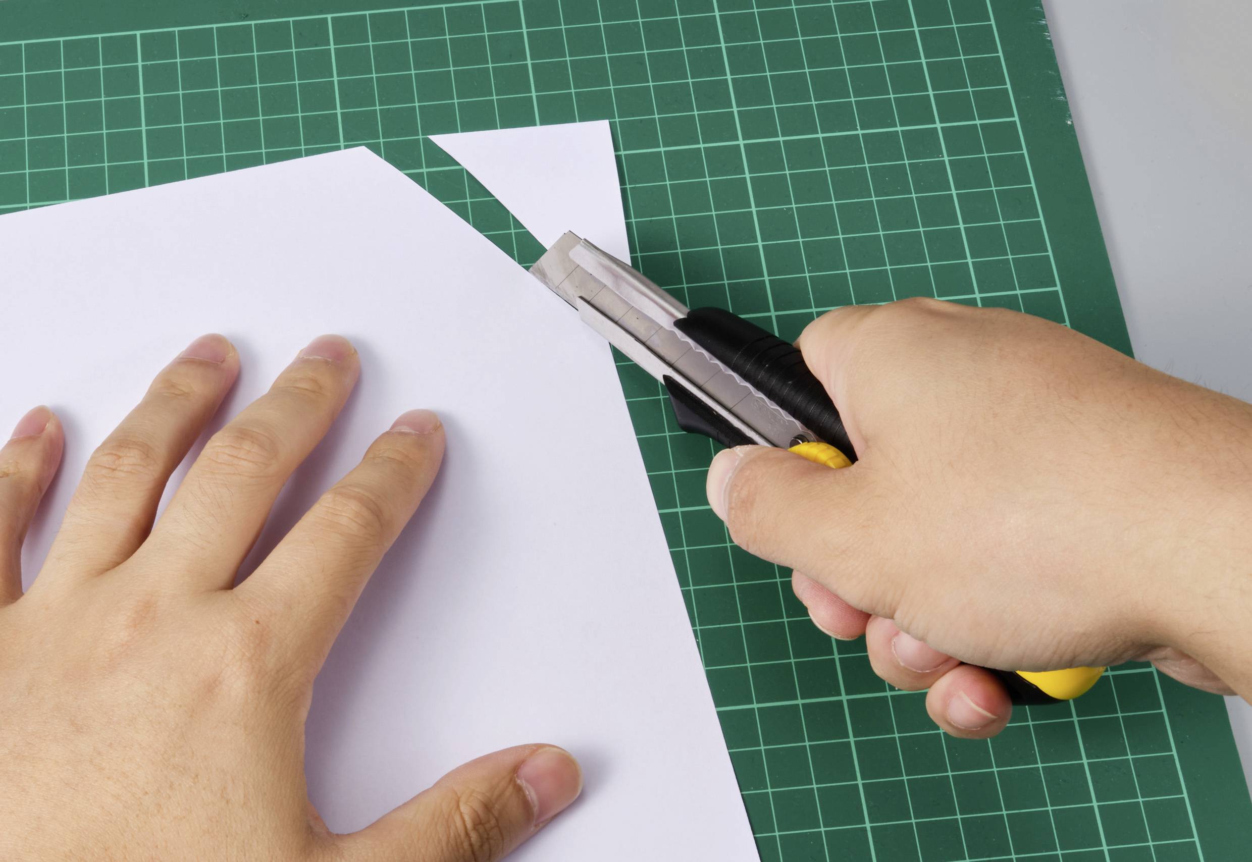 Eine Hand schneidet mit einem gelben Cutter-Messer ein weißes Papier auf einer grünen Schneidematte.