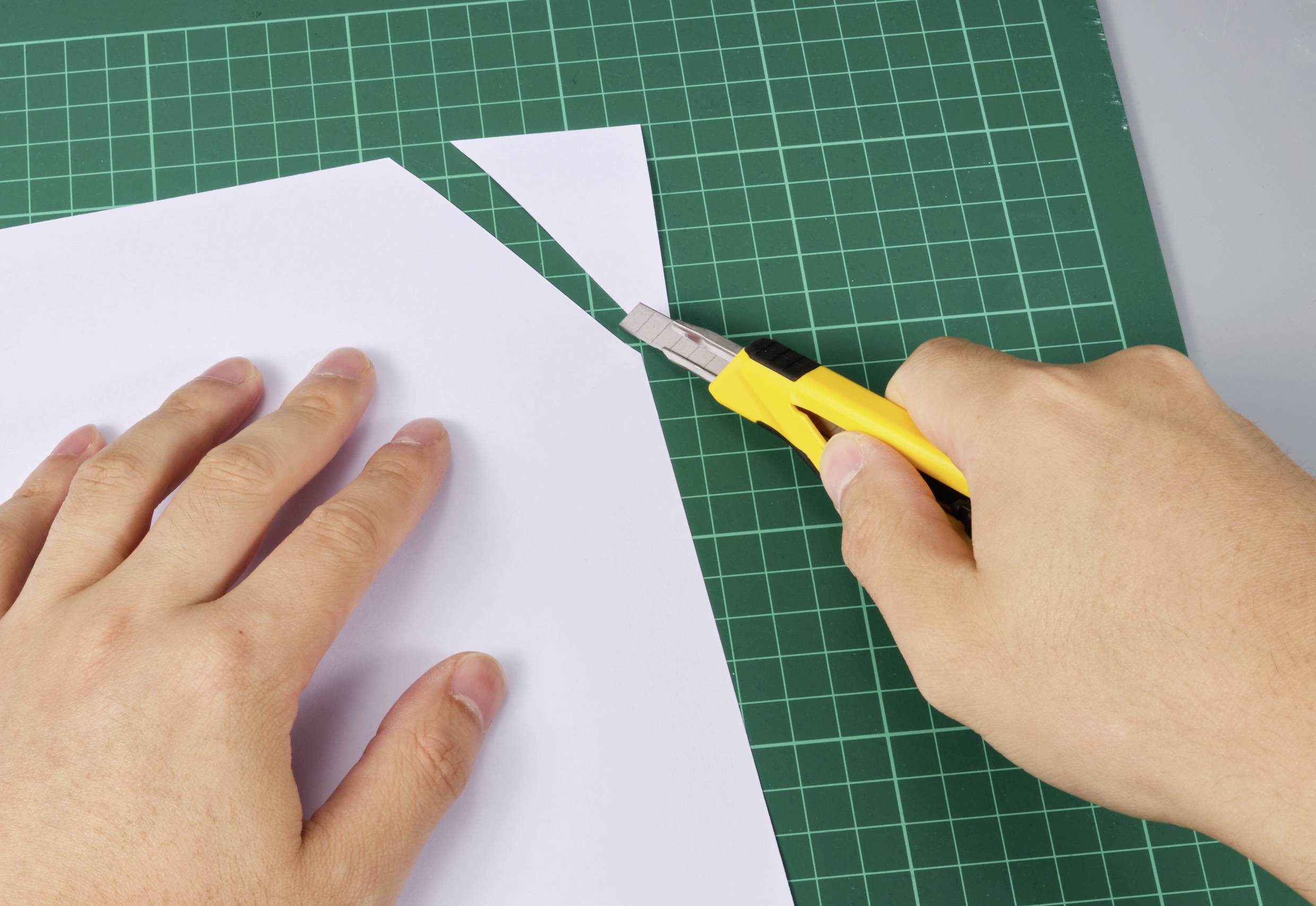 Eine Hand schneidet mit einem gelben Cuttermesser ein Dreieck aus einem weißen Blatt Papier auf einer grünen Schneidematte.