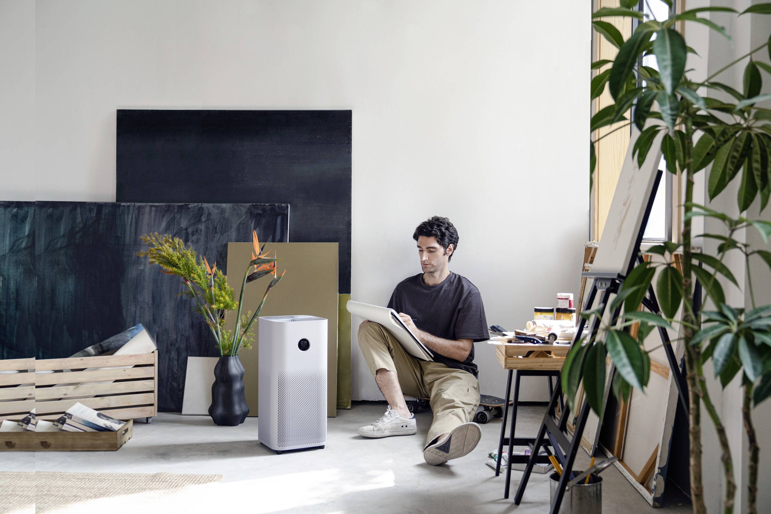 Ein Mann sitzt auf dem Boden eines hellen Raumes und malt auf einem Block. Neben ihm steht ein Luftreiniger und es gibt Pflanzen und Leinwände.