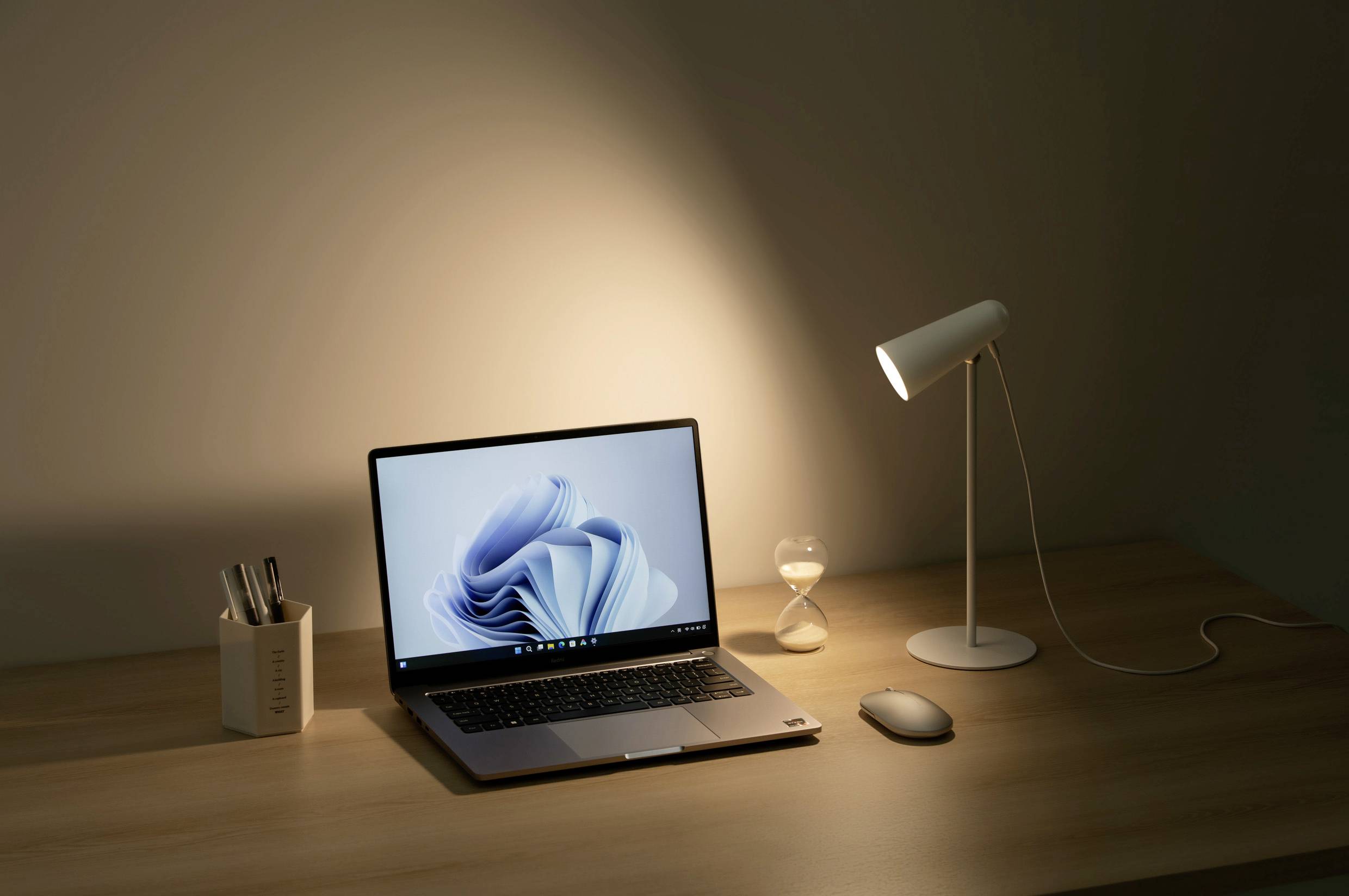 Ein beleuchteter Schreibtisch zeigt einen aufgeklappten Laptop, eine Sanduhr, eine Lampe und einen Stiftehalter unter sanftem Licht.