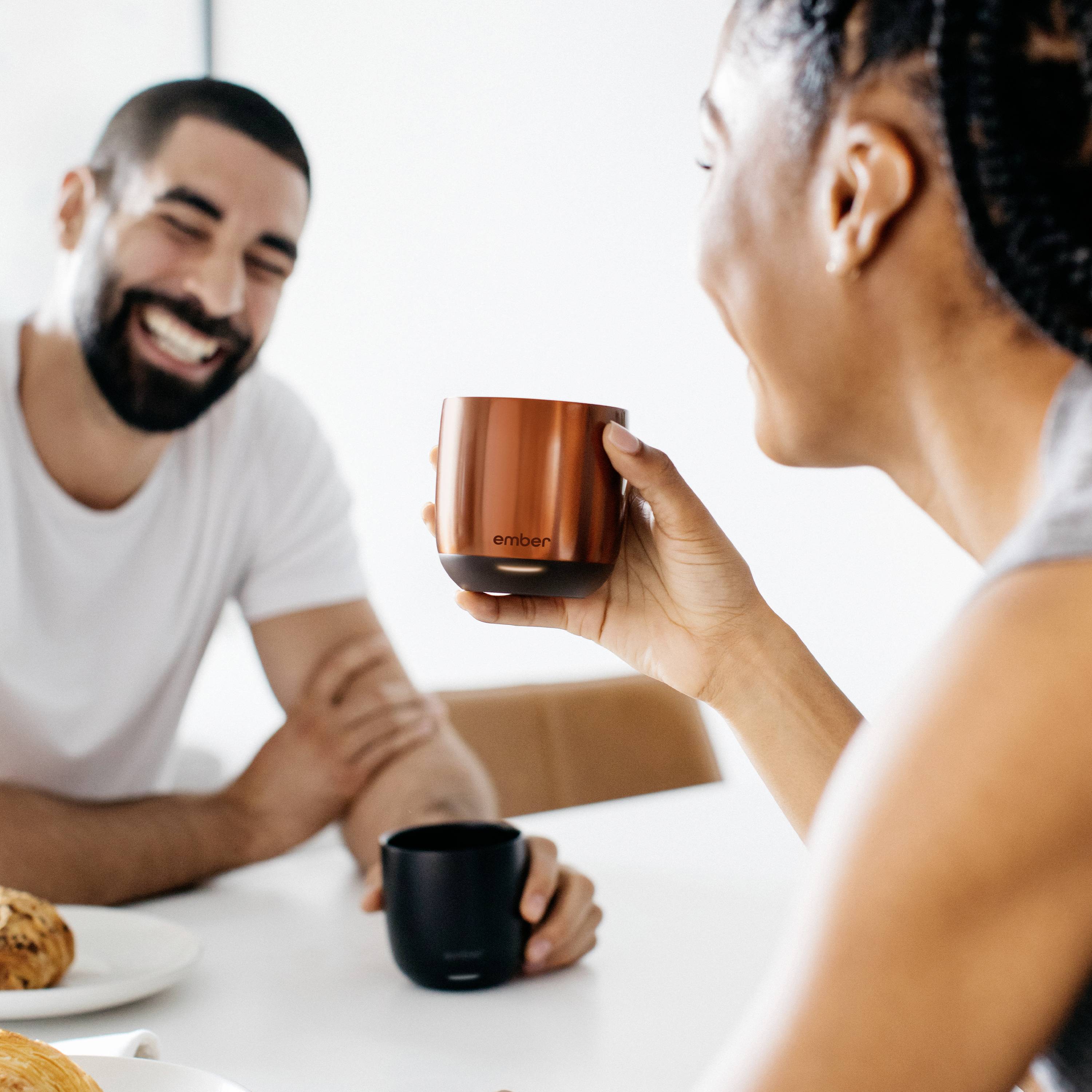 Eine Frau hält eine Kaffeetasse und lächelt zu einem Mann, der ebenfalls lächelt. Beide sitzen an einem Tisch mit Gebäck und Kaffee.
