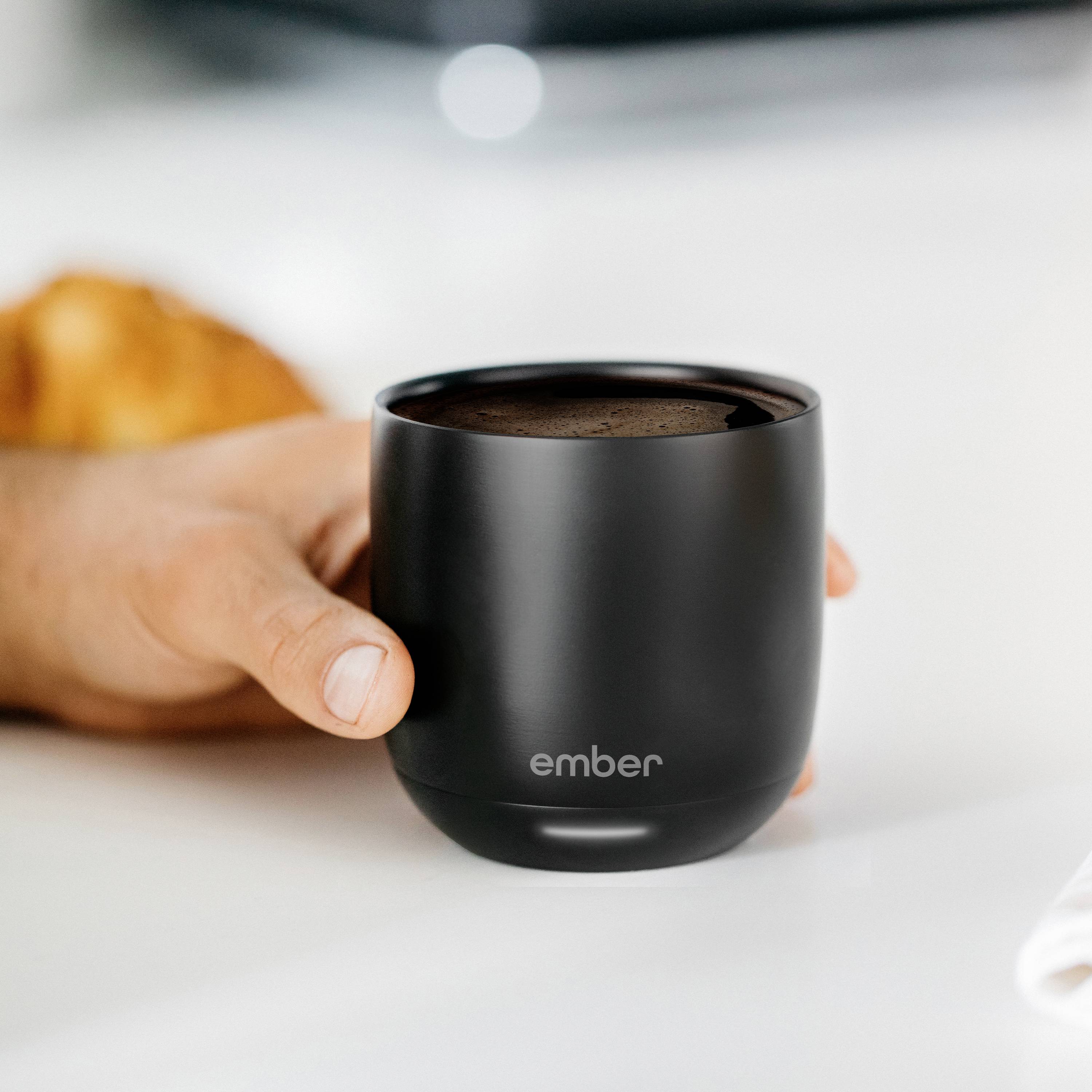 Eine Hand hält eine schwarze, beheizbare Tasse mit der Aufschrift 'ember'. Im Hintergrund ist ein verschwommener Gegenstand zu sehen.
