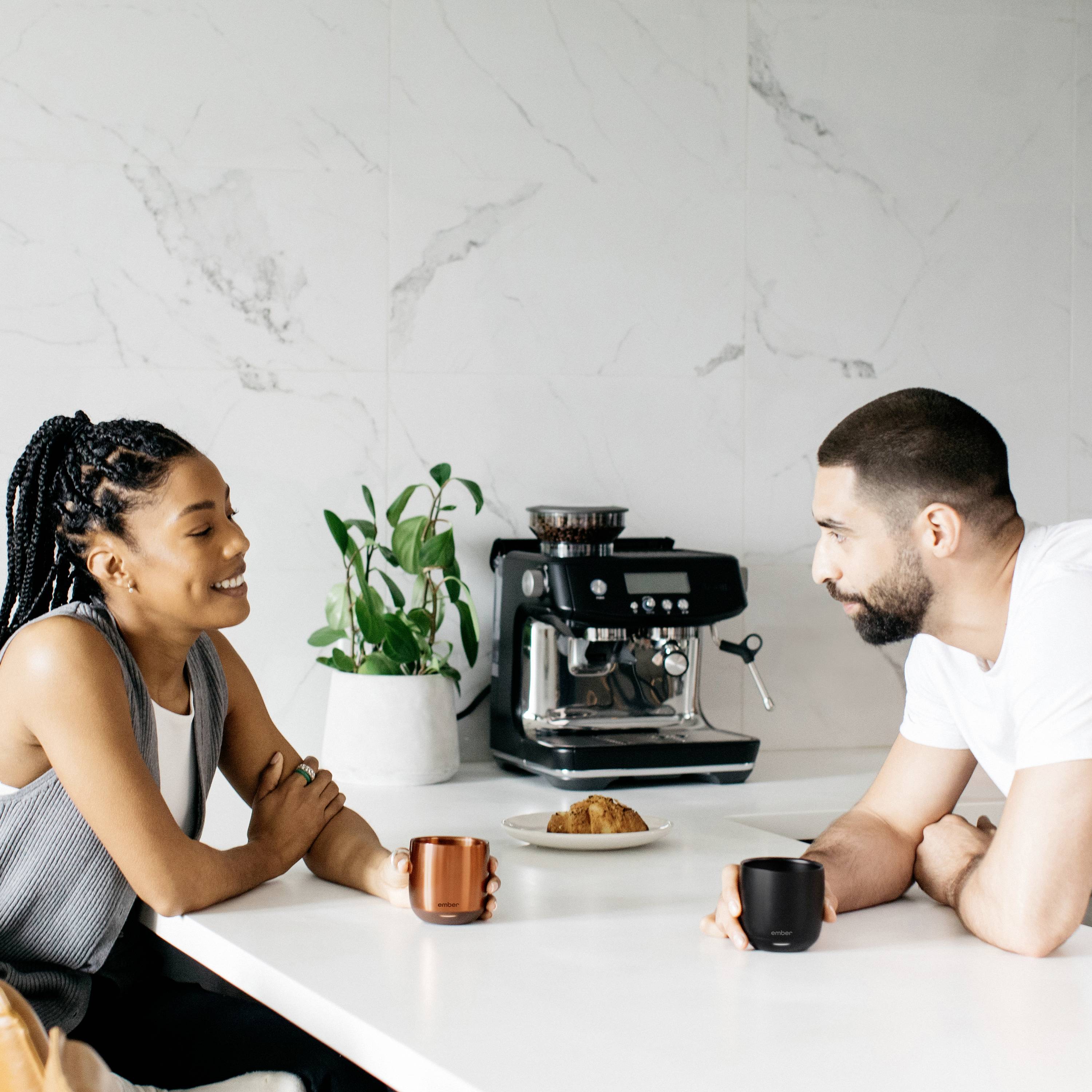 Eine Frau und ein Mann sitzen an einem weißen Küchentresen, trinken Kaffee und unterhalten sich. Im Hintergrund steht eine Kaffeemaschine.