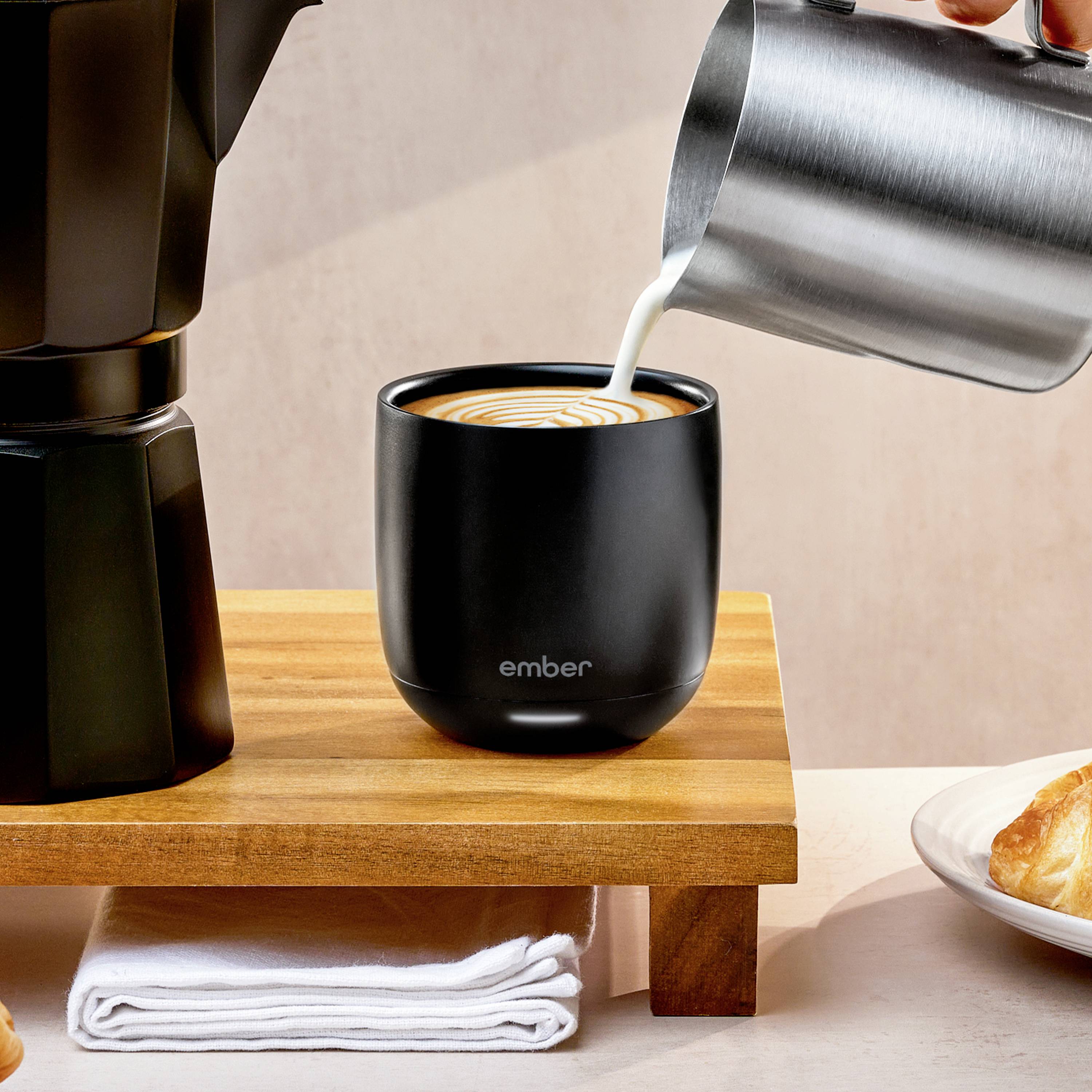Eine silberne Kanne gießt Milch in eine schwarze Tasse auf einem Holztisch. Daneben steht eine Mokkakanne.