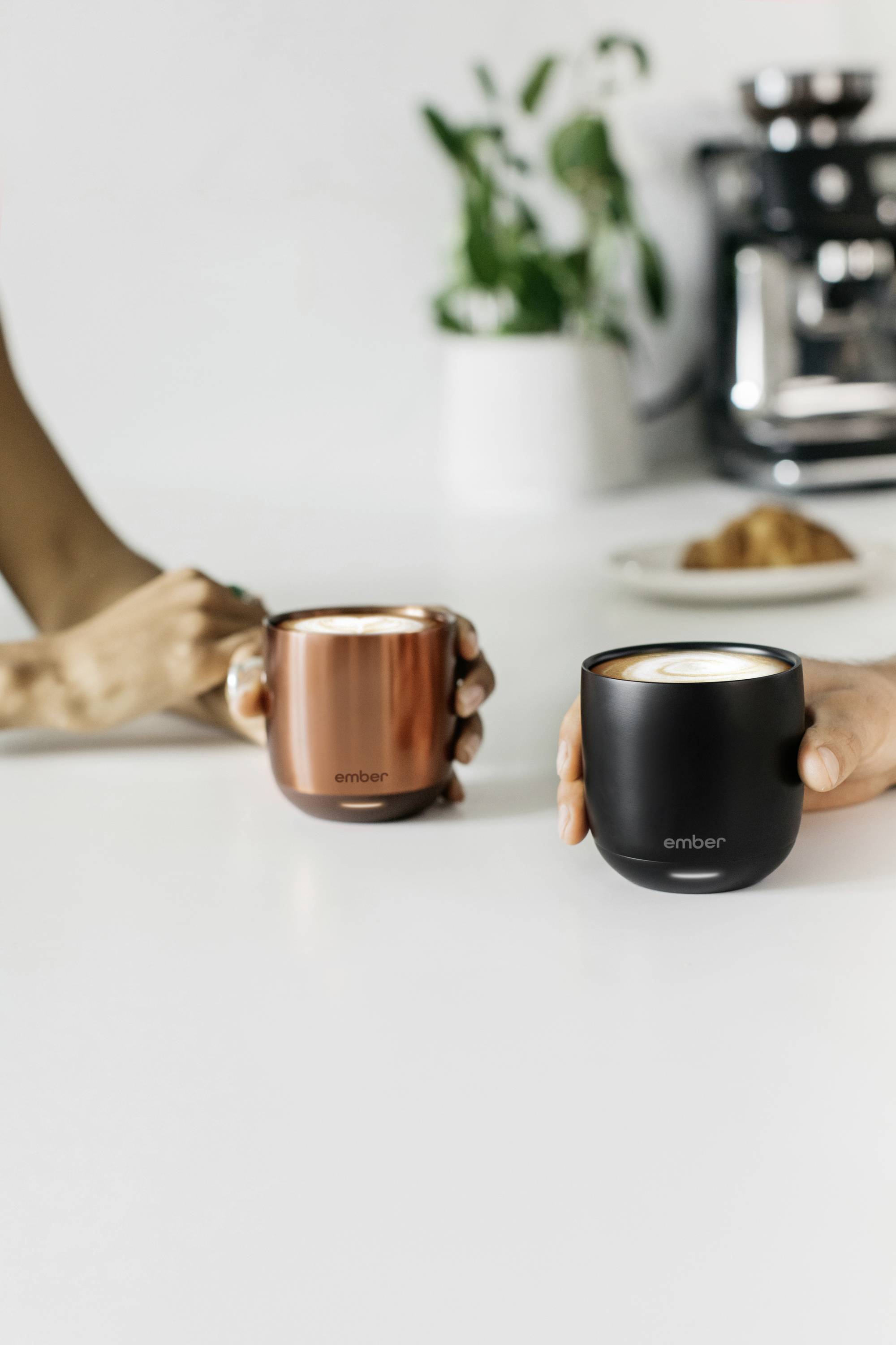 Zwei Hände halten metallische Tassen mit Kaffee. Eine Pflanze und ein Kuchen sind im Hintergrund auf einem weißen Tisch.