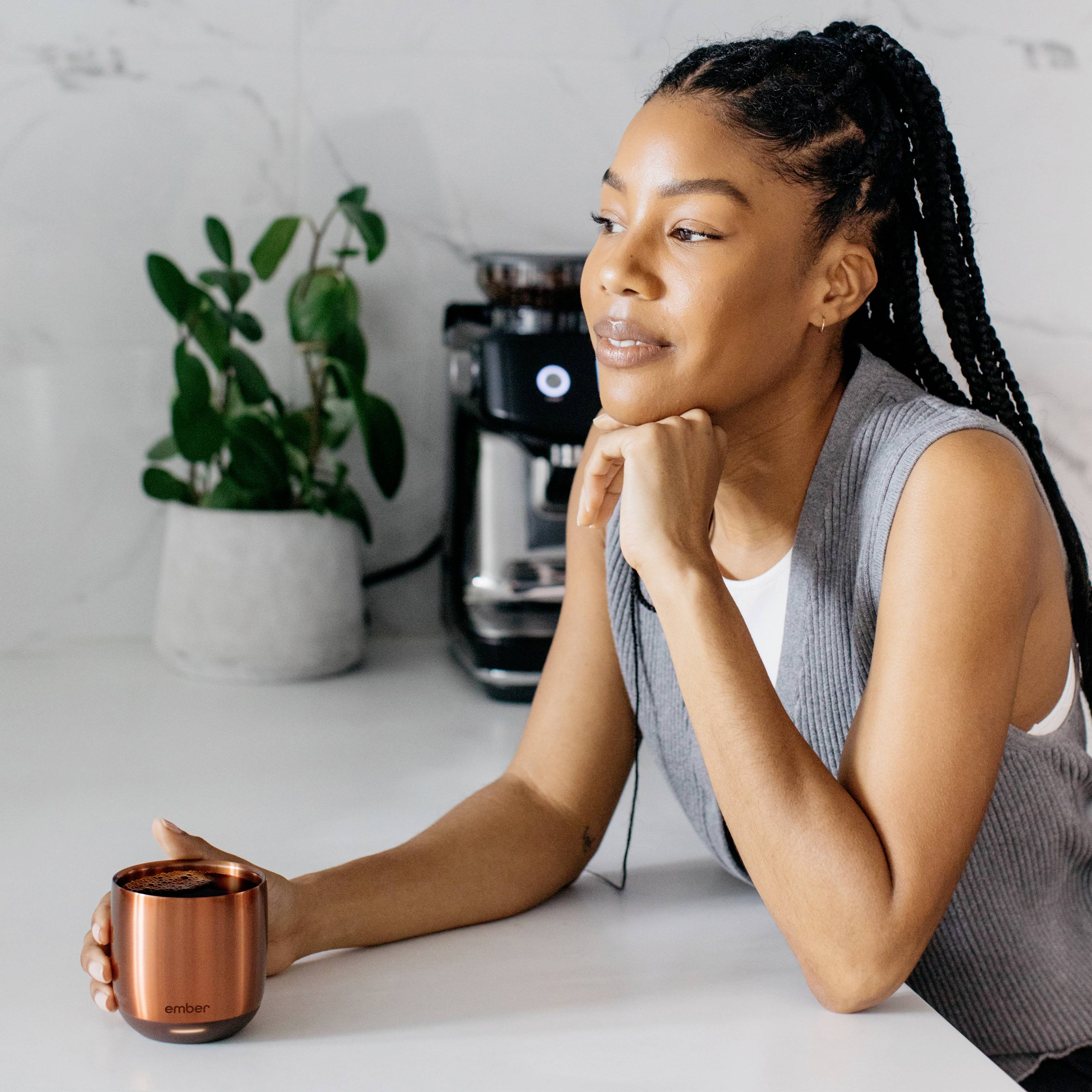 Eine Frau sitzt am Tisch mit einer Kupfertasse in der Hand. Im Hintergrund sind eine Pflanze und eine Kaffeemaschine zu sehen.