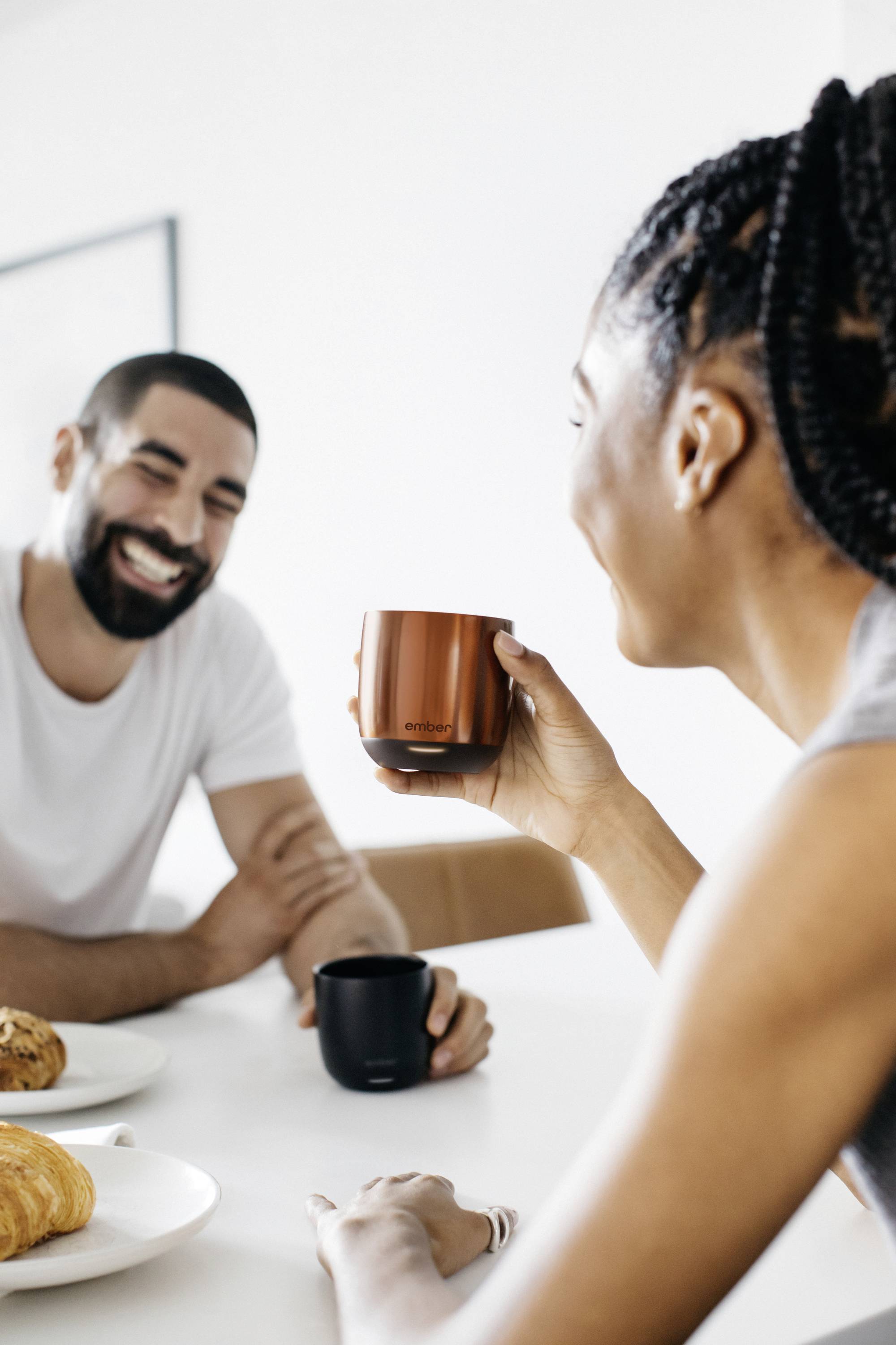 Zwei Personen sitzen an einem Tisch, trinken aus Tassen und lachen. Auf dem Tisch liegen Croissants. Entspannte Frühstücksszene.