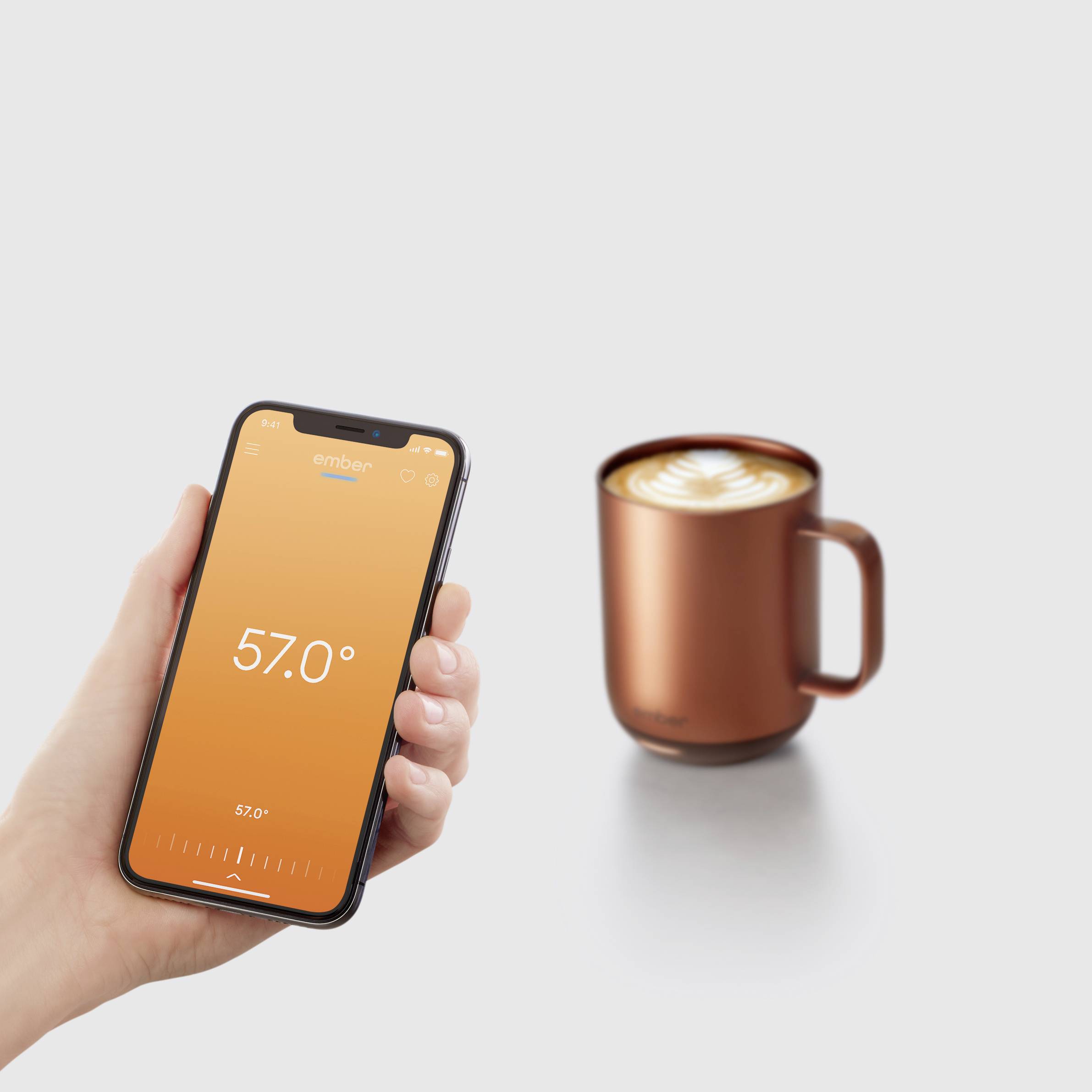 Eine Hand hält ein Smartphone mit einer App, die die Temperatur von 57,0 Grad anzeigt. Daneben steht eine Tasse Kaffee mit Latte Art.