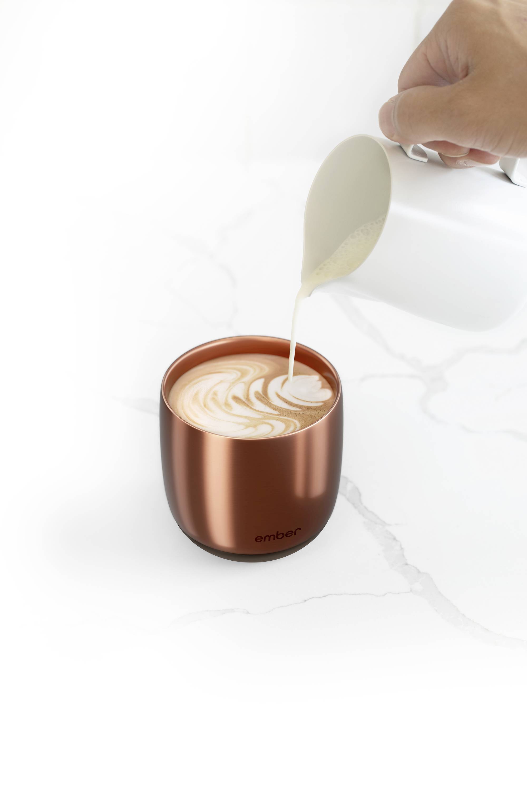 Eine Hand gießt Milch in eine Tasse mit Latte Art. Die Tasse steht auf einer weißen Marmoroberfläche.