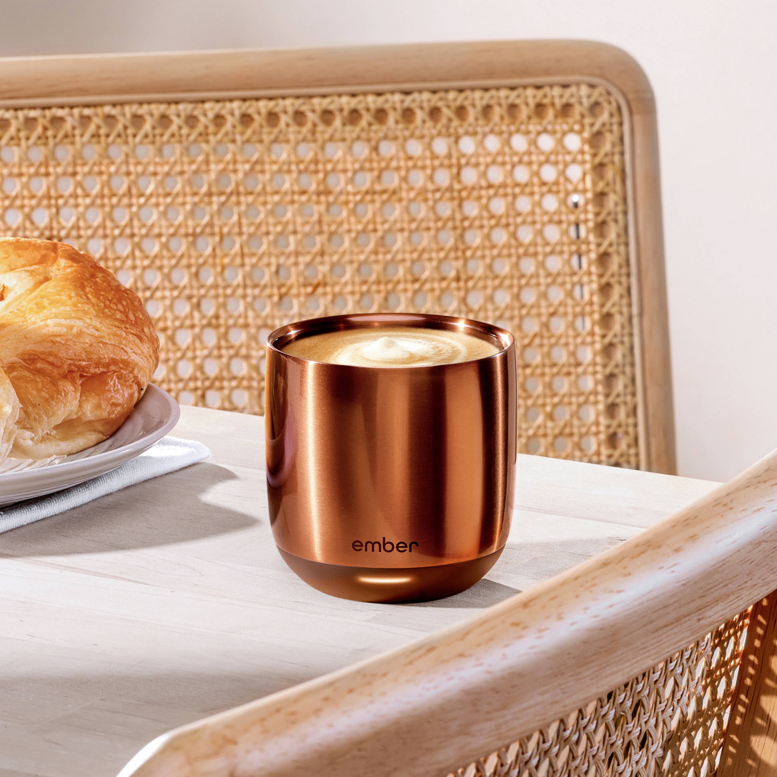 Ein Kaffeebecher mit Schaum, auf einem Tisch neben einem Croissant. Der Becher hat ein kupferfarbenes Design mit dem Logo 'ember'.