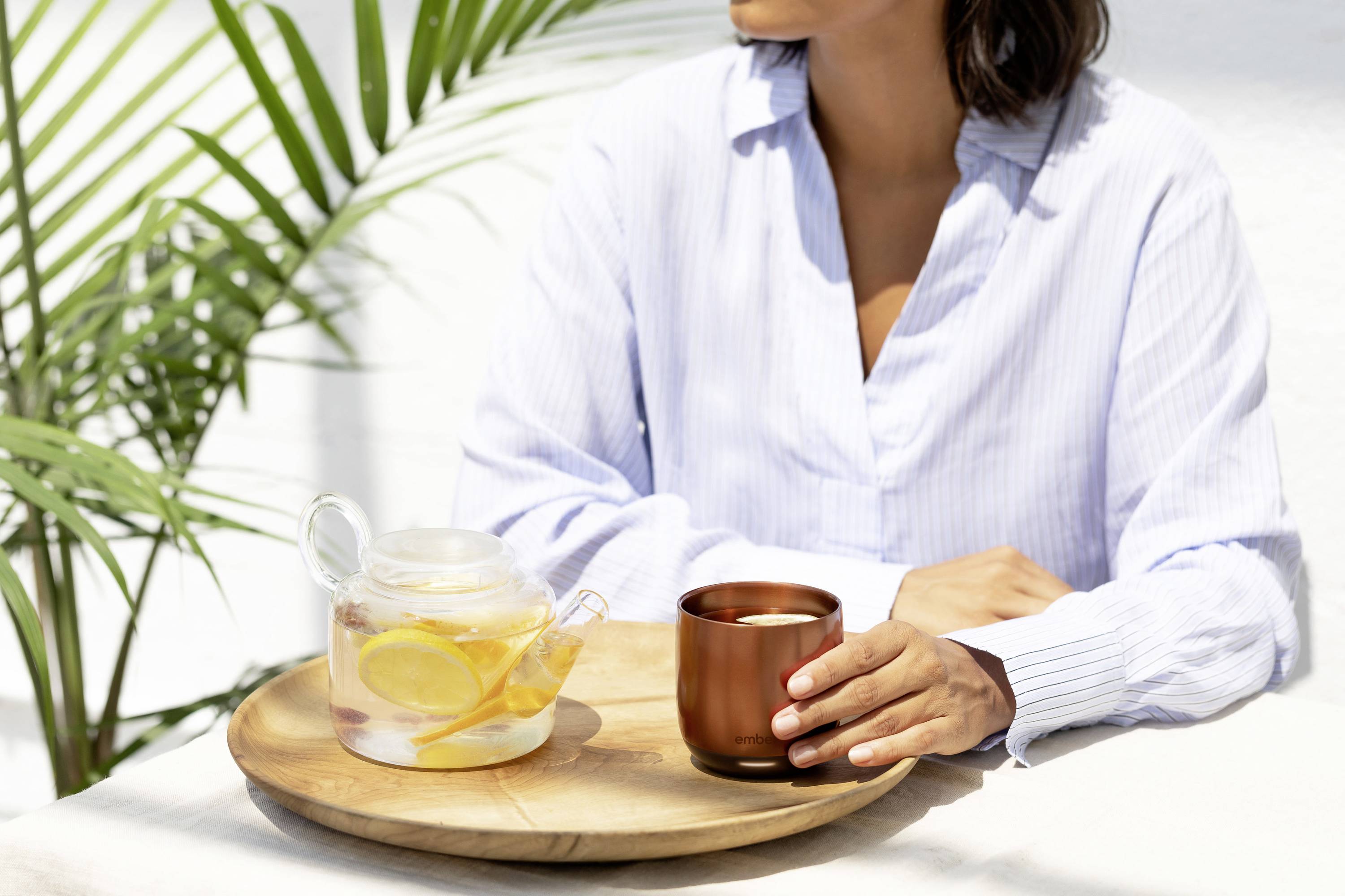Eine Person sitzt an einem Tisch und hält eine Tasse. Daneben steht eine Karaffe mit Zitronenwasser. Im Hintergrund sind Pflanzen sichtbar.