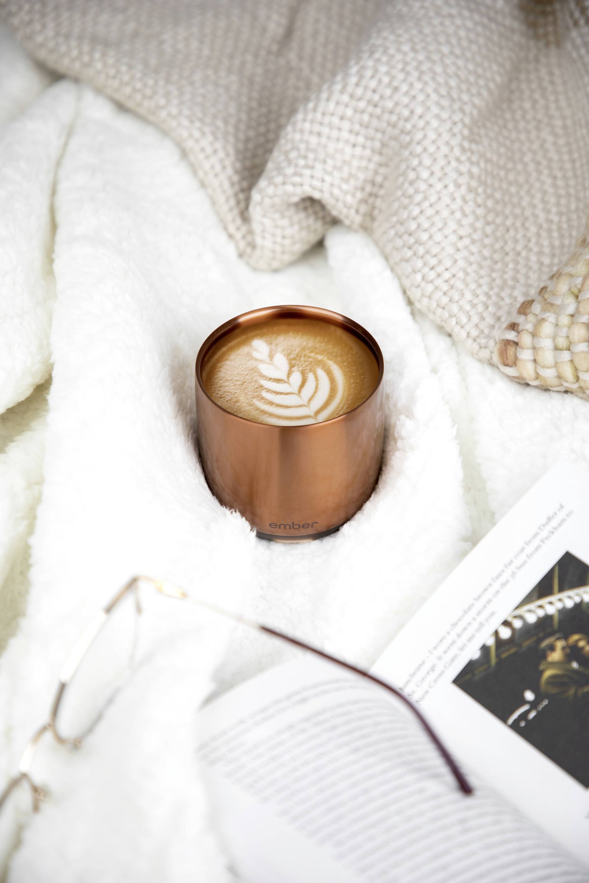 Eine Tasse Cappuccino mit Blatt-Latte-Art auf einem weißen, flauschigen Untergrund neben einem aufgeschlagenen Buch und einer Brille.