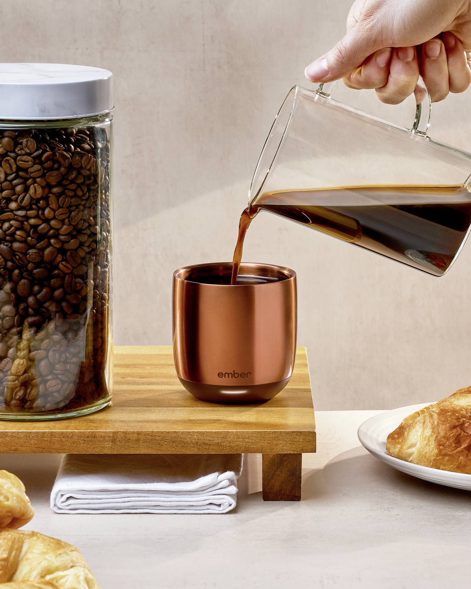 Eine Hand gießt Kaffee aus einer Glaskanne in eine kupferfarbene Tasse. Daneben stehen ein Glas mit Kaffeebohnen und Gebäck.