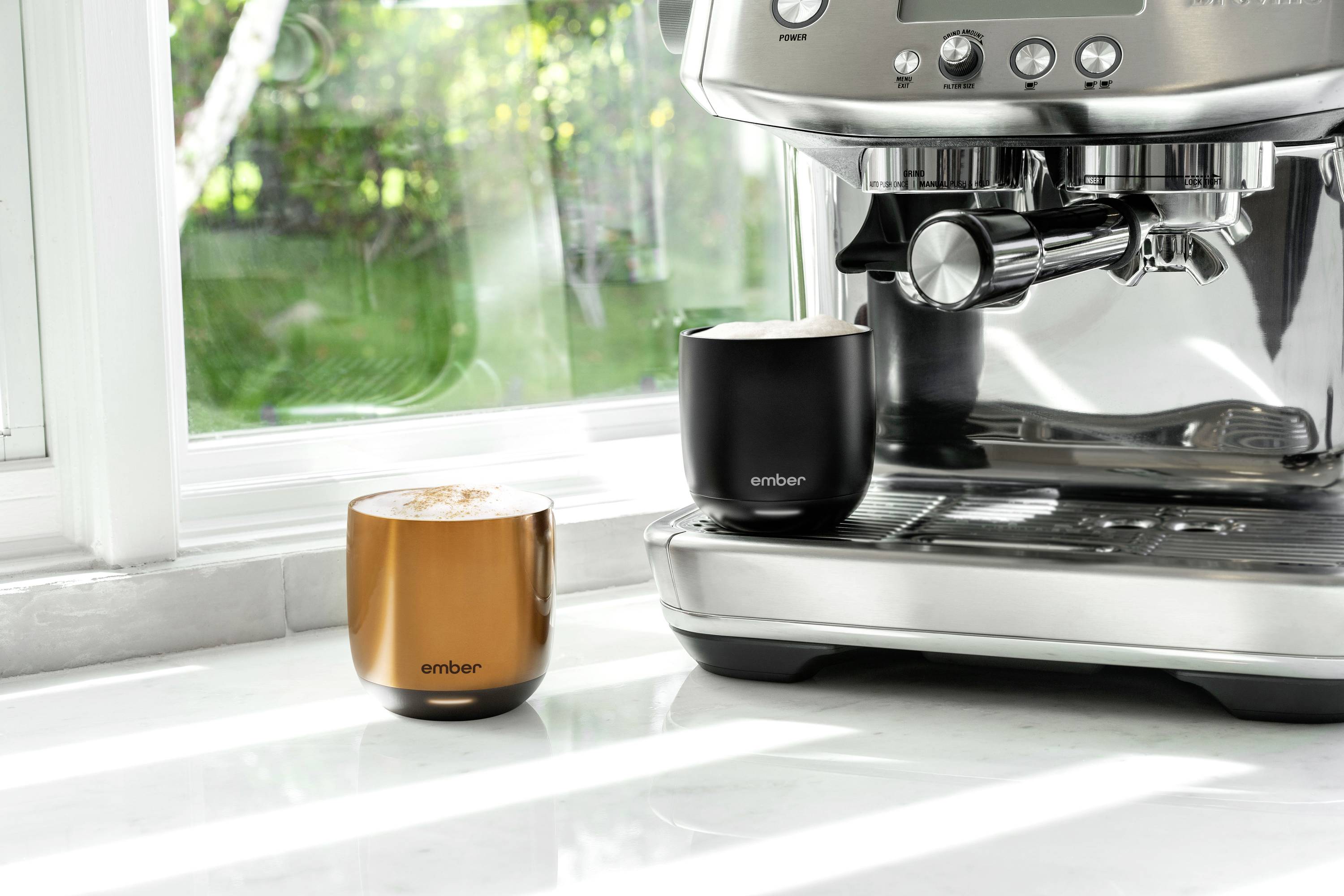 Elektrische Kaffeemaschine auf einer Arbeitsfläche neben zwei Kaffeebechern, einer in Bronze und einer in Schwarz, vor einem Fenster.