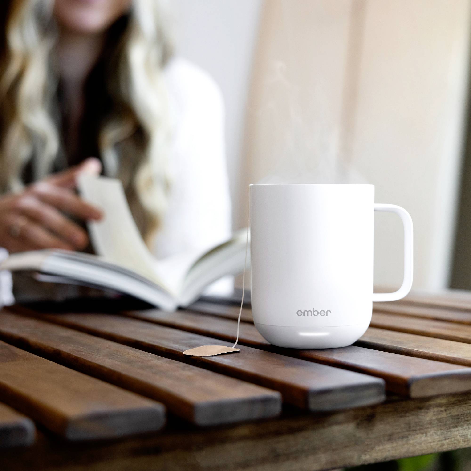 Eine weiße Tasse, aus der Dampf aufsteigt, steht auf einem Holztisch. Im Hintergrund liest eine Person ein Buch.