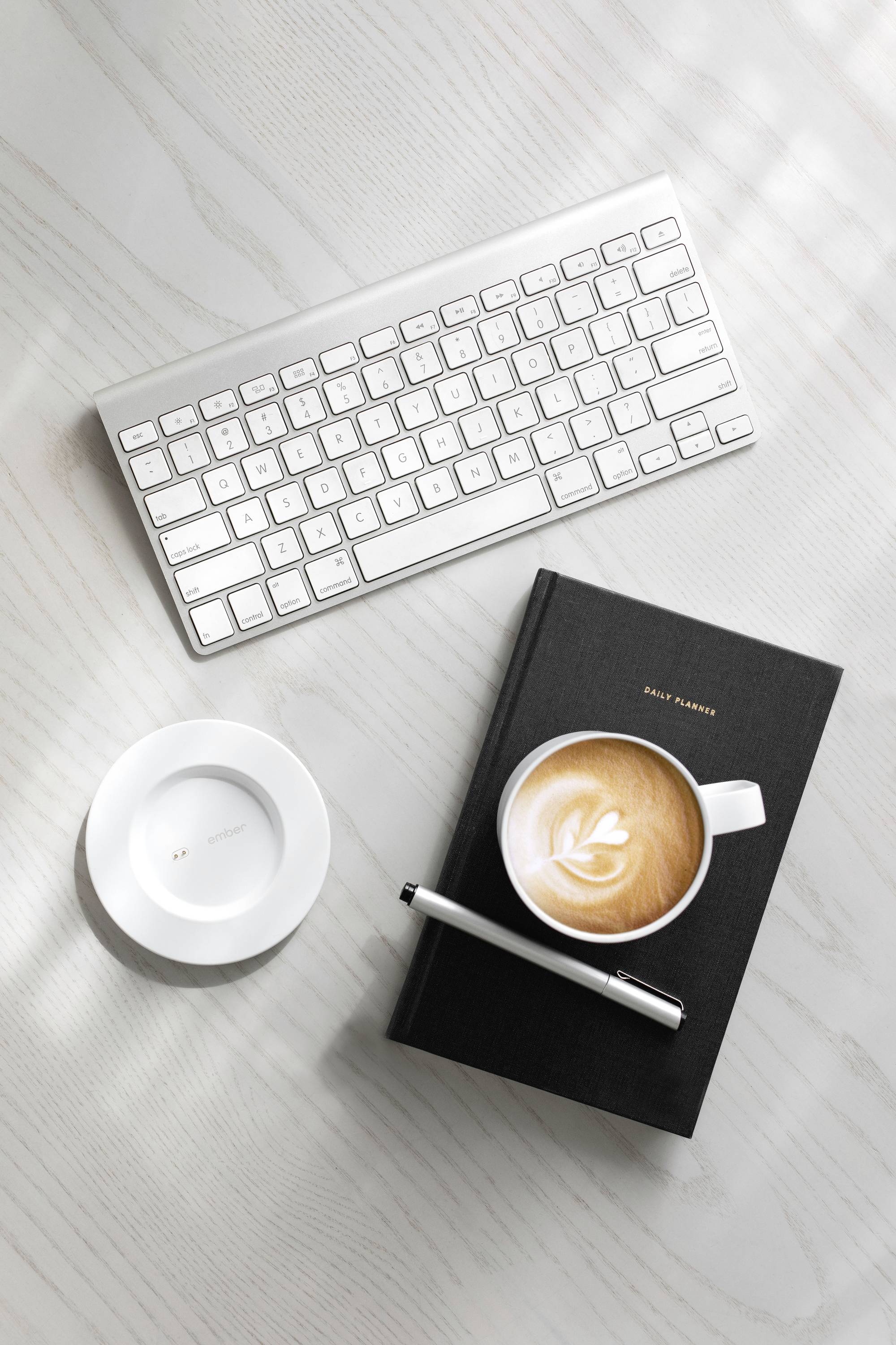Tastatur, Notizbuch mit Stift und Tasse Cappuccino auf einem Schreibtisch. Büroarbeitsplatz mit Getränk und Schreibmaterialien.