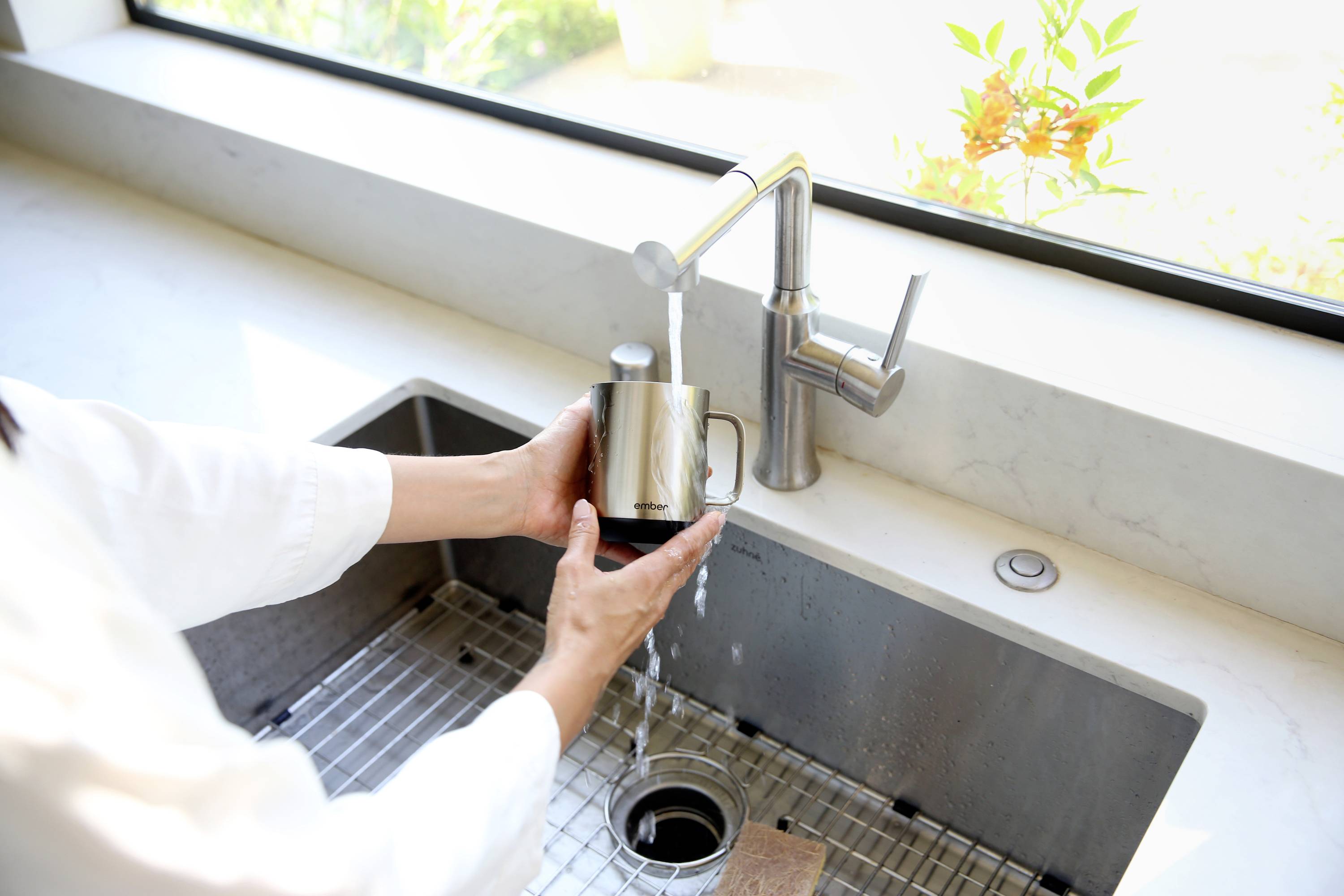 Person füllt eine Metalltasse mit Wasser aus einem Küchenhahn am Spülbecken. Helles Tageslicht fällt durch das Fenster dahinter.