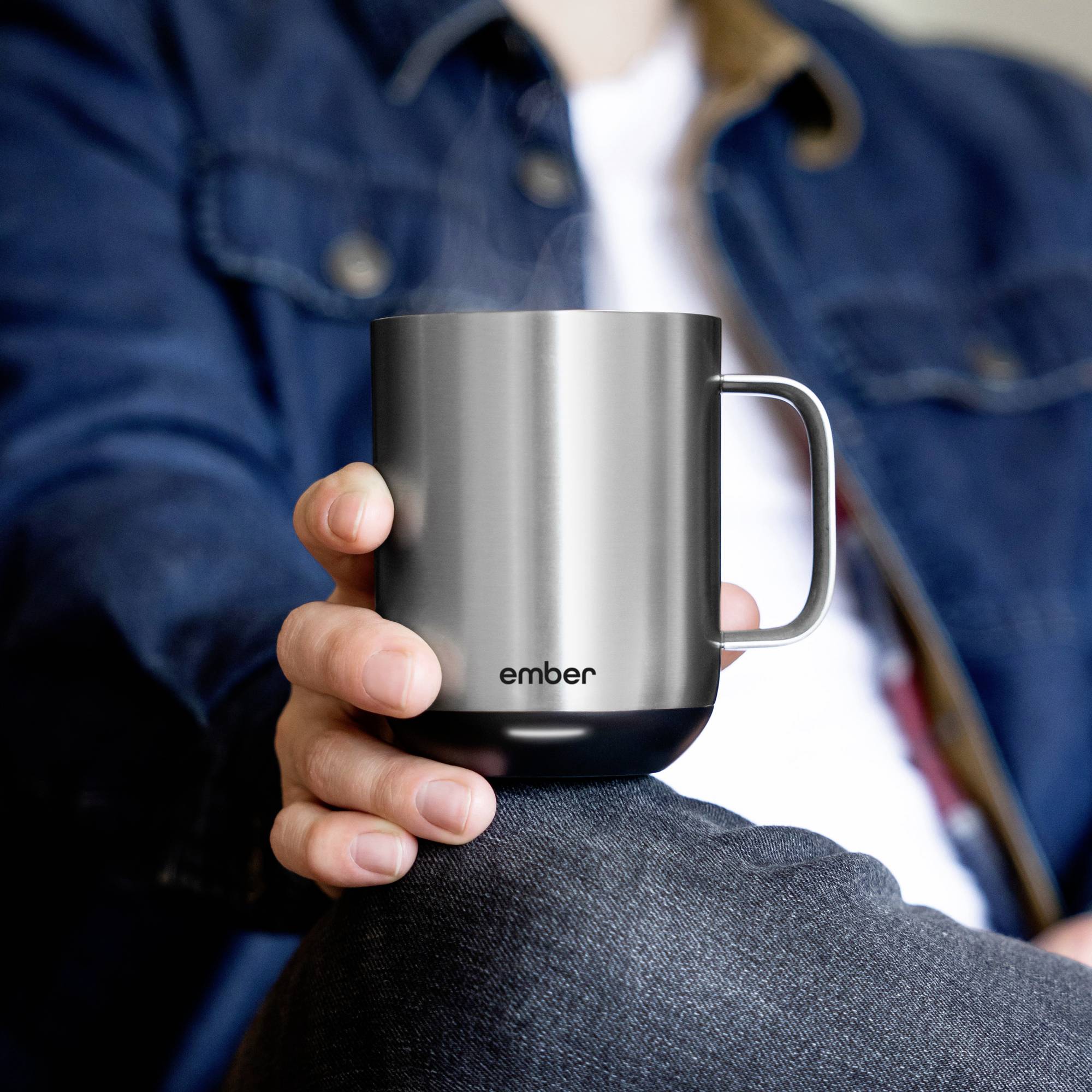 Eine Person hält eine graue Tasse mit dem Schriftzug 'ember', aus der Dampf aufsteigt, in ihrer Hand. Die Person trägt eine dunkelblaue Jacke.