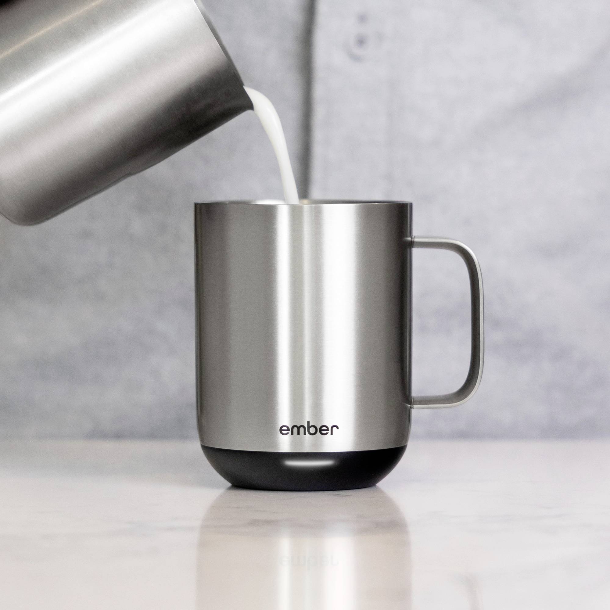Eine Edelstahl-Tasse mit der Aufschrift 'ember' wird mit Milch aufgefüllt. Die Tasse steht auf einer grauen Oberfläche.