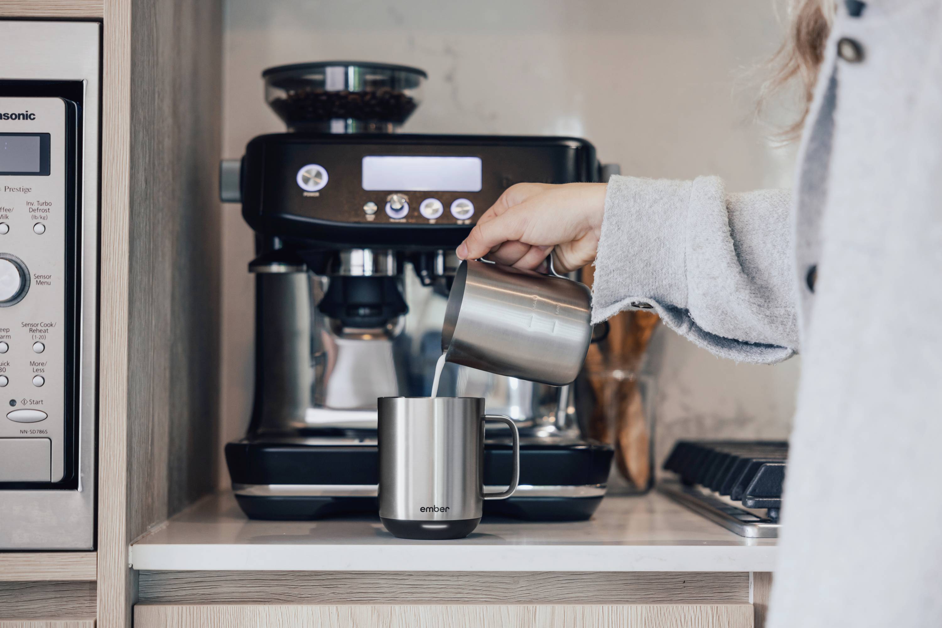 Eine Person gießt Milch in eine Tasse vor einer Espressomaschine in einer Küche. Im Hintergrund sind Küchengeräte sichtbar.