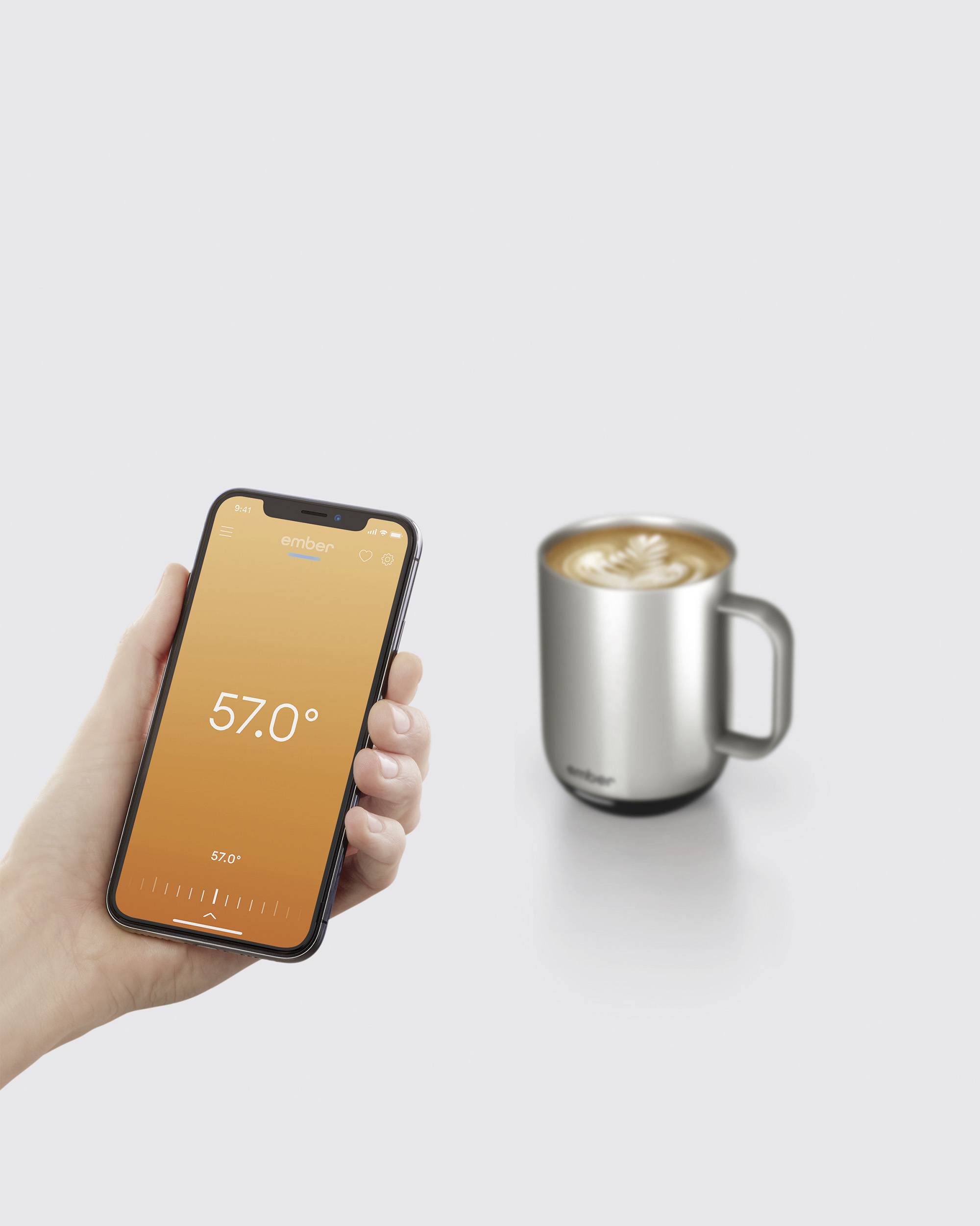 Eine Hand hält ein Smartphone, das eine Temperatur von 57 Grad anzeigt. Daneben steht ein silberner Becher mit einer heißen Getränk.