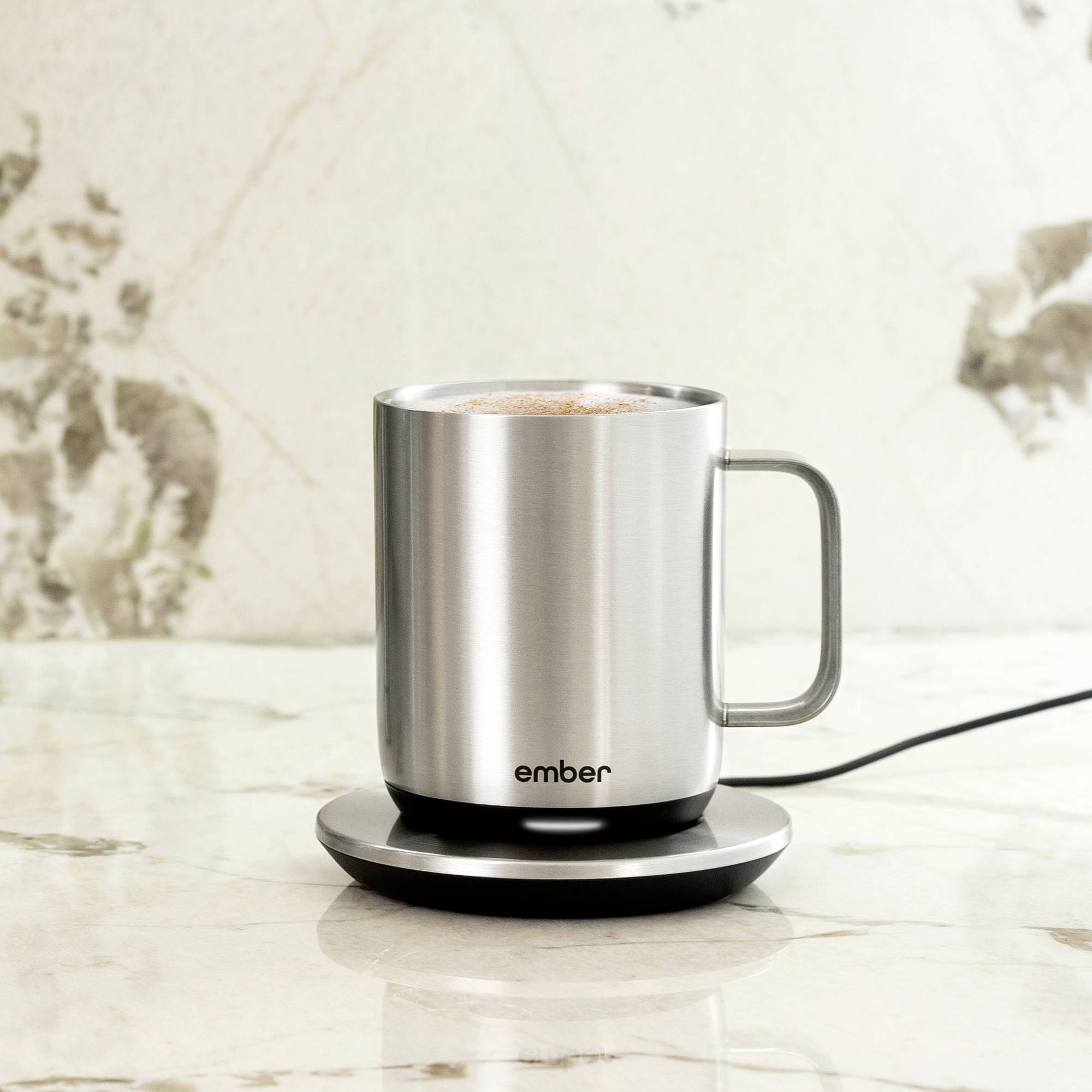 Eine silberne Tasse mit 'ember'-Aufdruck steht auf einer Ladestation, gefüllt mit einem heißen Getränk. Sie befindet sich auf einer Marmoroberfläche.