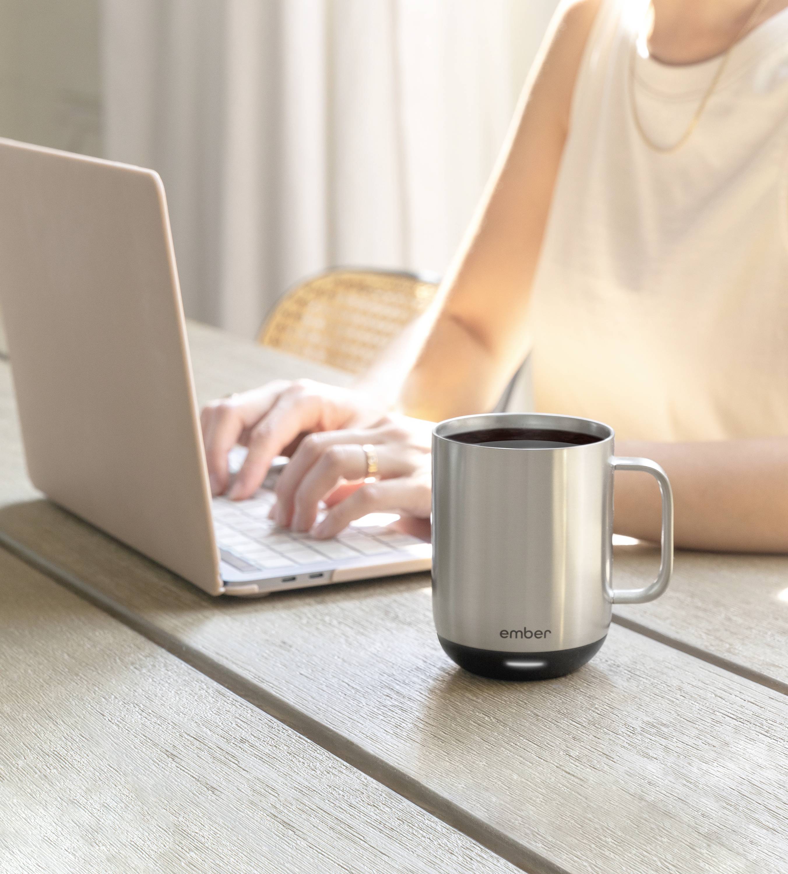 Eine Person tippt auf einem Laptop an einem Tisch, neben einem silbernen Thermobecher.