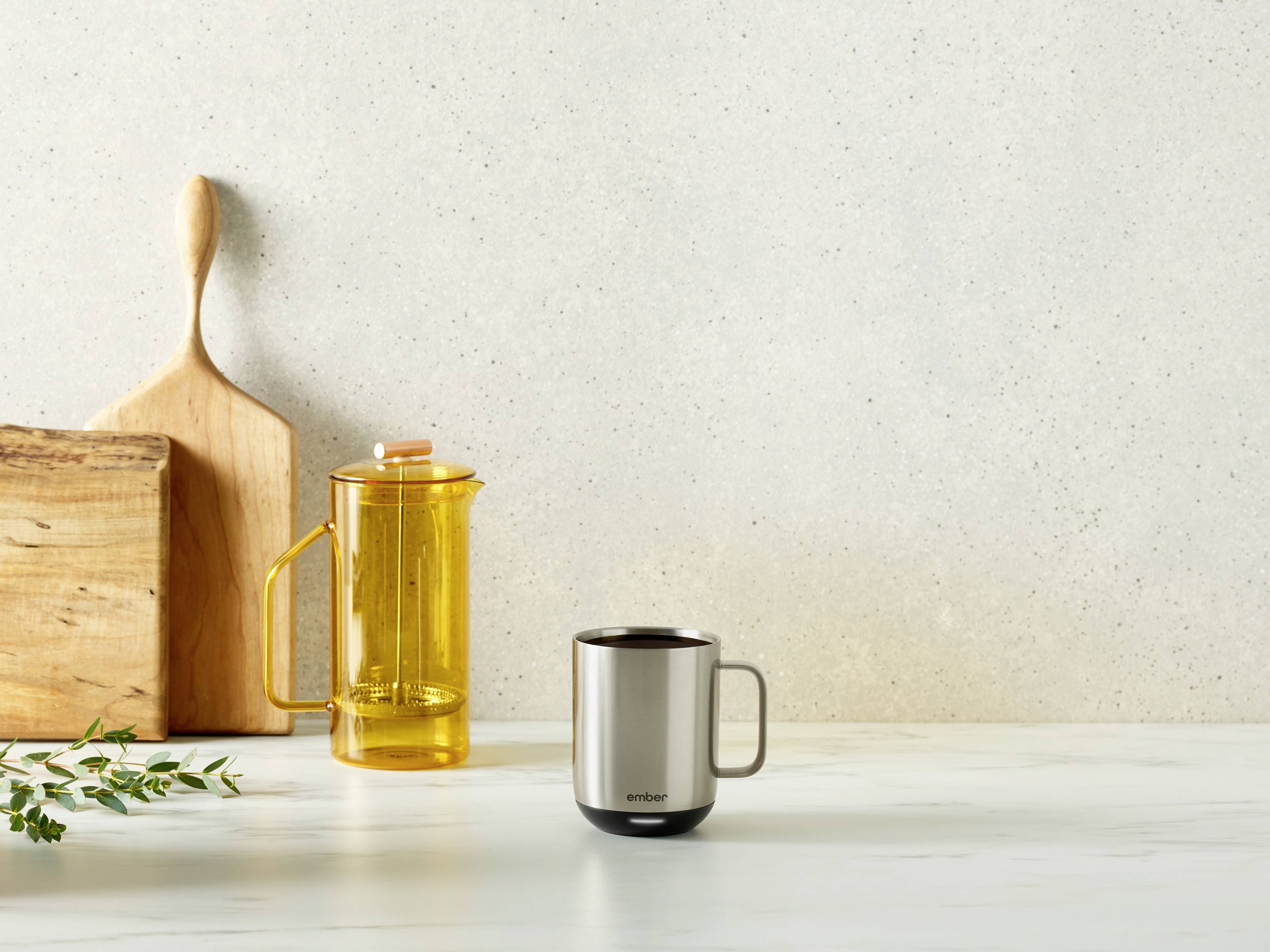 Eine Metalltasse auf einem Marmortisch, daneben eine gelbe Teekanne und ein Holzschneidebrett vor einem grauen Hintergrund.