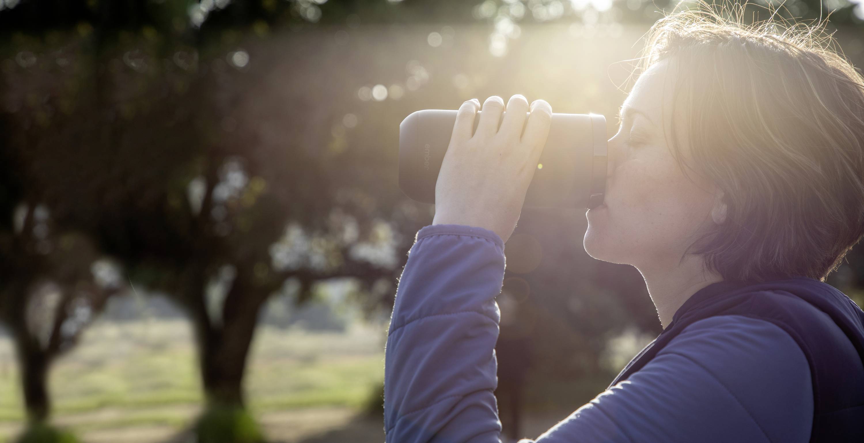 Eine Person in Freizeitkleidung trinkt aus einer Thermoskanne im Freien, während Sonnenlicht durch Bäume scheint.