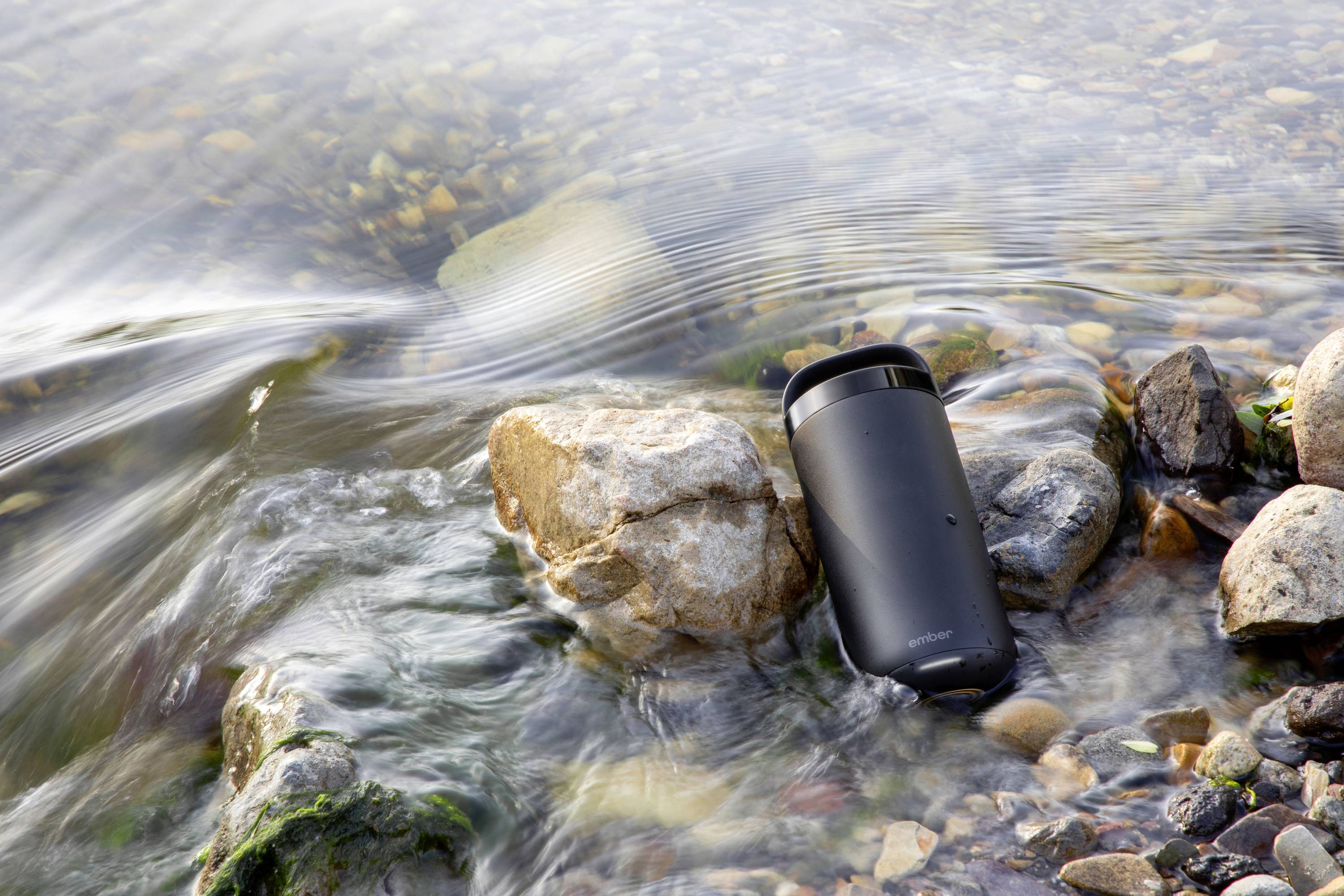 Schwarzer Thermobecher liegt in fließendem Wasser zwischen Steinen am Ufer.