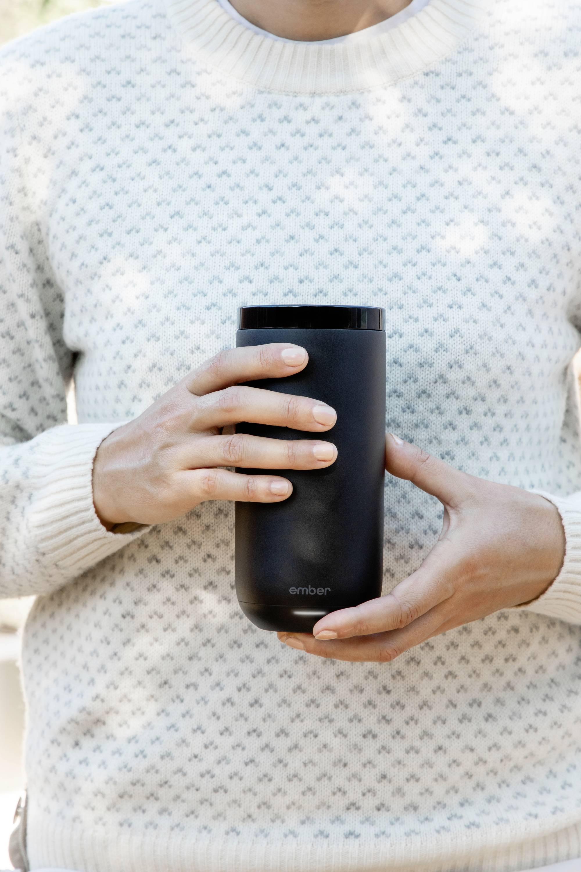 Person hält schwarzen Thermobecher in beiden Händen. Hintergrund unscharf, Fokus auf Becher.