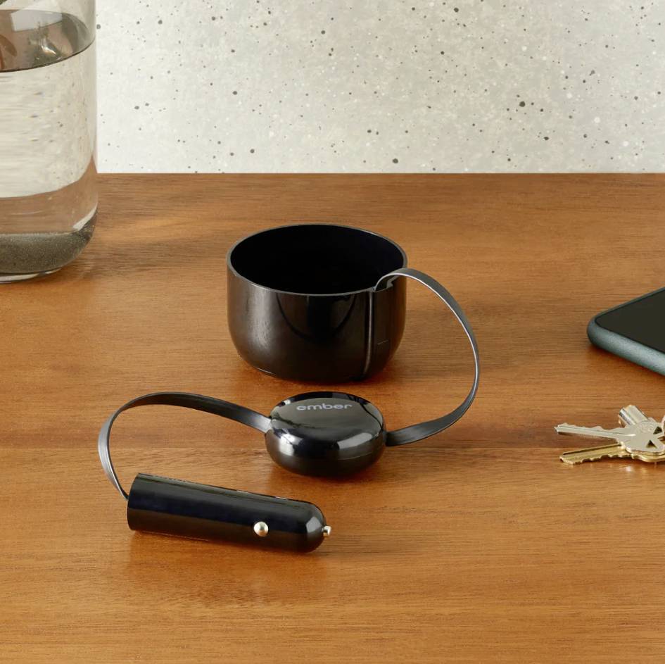 Eine schwarze, temperaturgeregelte Tasse auf einem Tisch, daneben ein Smartphone, Schlüsselbund und ein Glas Wasser.