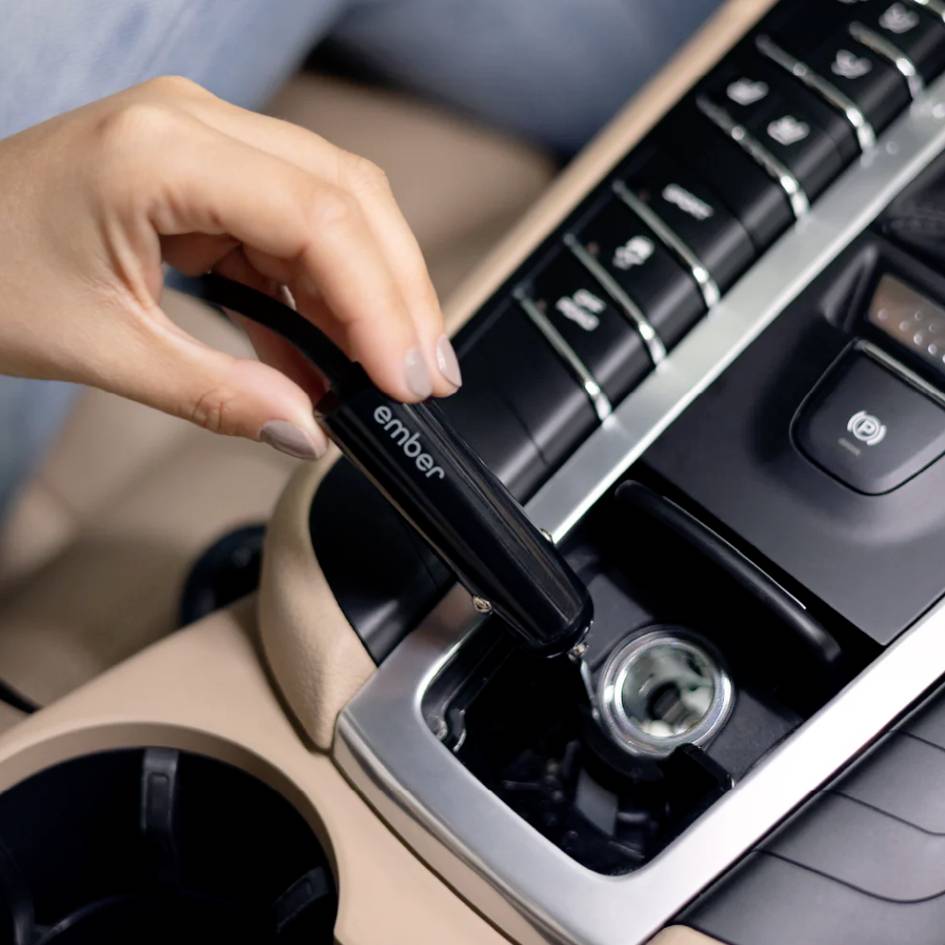 Eine Hand hält einen 'ember'-Adapter und steckt ihn in einen Auto-Zigarettenanzünder. Das Armaturenbrett des Autos ist sichtbar.