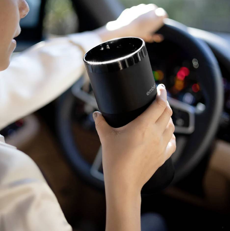 Person hält einen schwarzen Thermobecher in der Hand, während sie ein Auto steuert. Im Hintergrund ist das Lenkrad sichtbar.