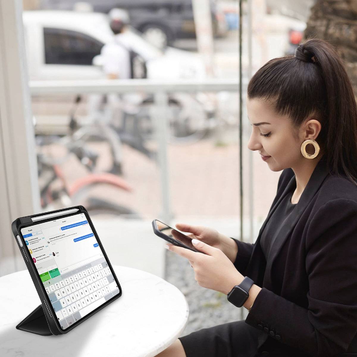 Eine Frau sitzt in einem Café und schaut auf ihr Smartphone. Neben ihr steht ein Tablet auf einem Tisch, das eine Chat-Anwendung zeigt.