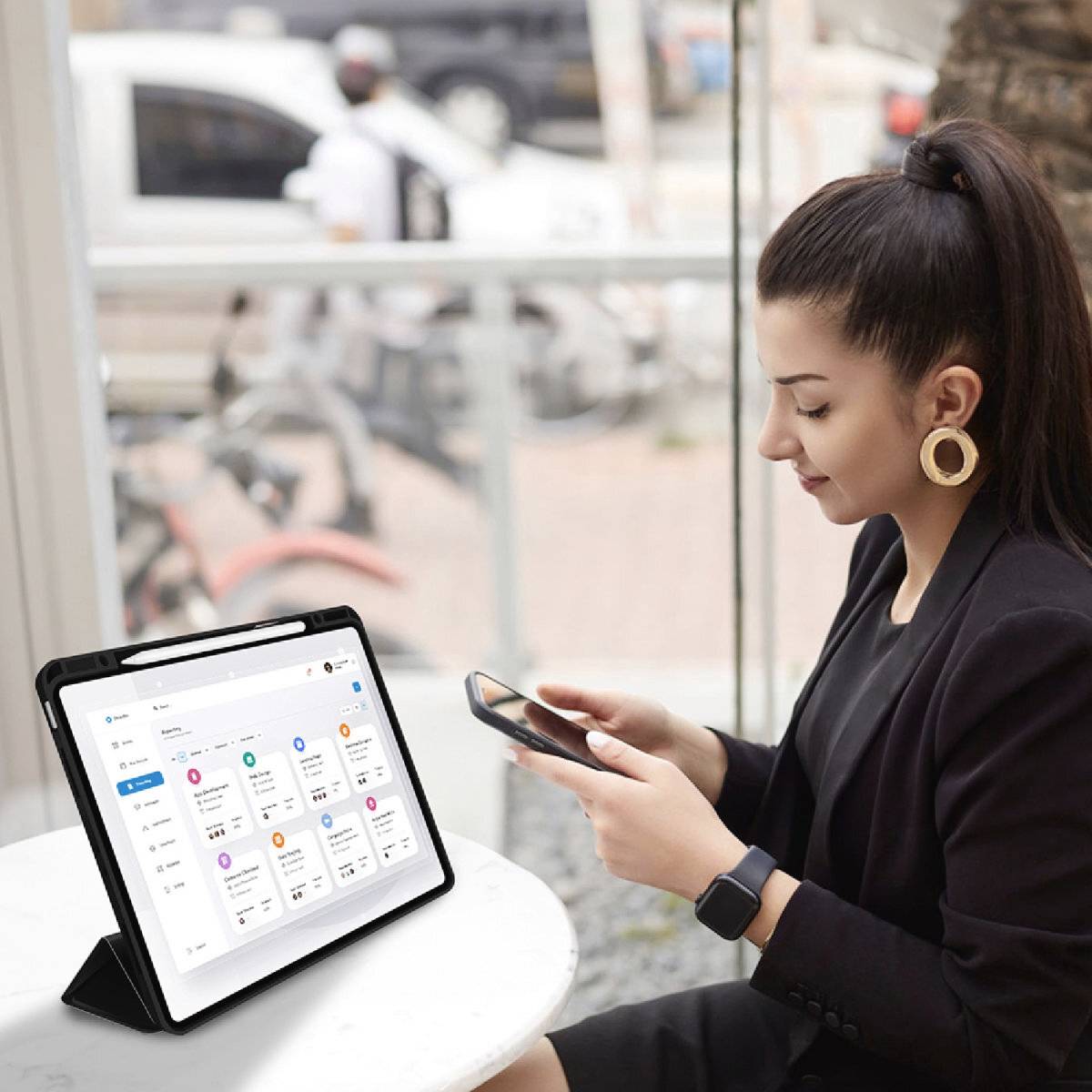 Frau mit Smartphone sitzt an einem Tisch, neben ihr ein Tablet mit geöffnetem Kalender. Sie wirkt konzentriert.