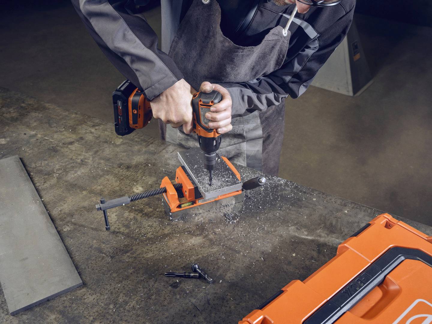 Eine Person arbeitet mit einer Bohrmaschine an einem Metallstück, das in einem Schraubstock auf einem Arbeitstisch eingespannt ist.