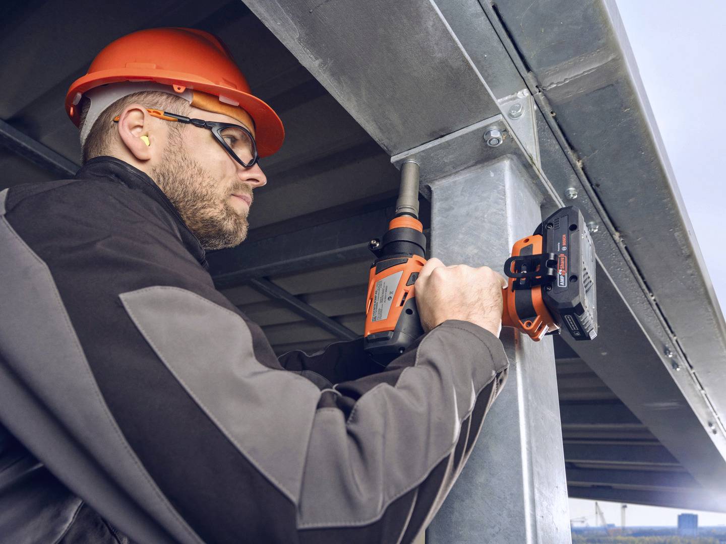 Ein Bauarbeiter mit Schutzhelm und Brille verwendet eine Bohrmaschine, um Metallträger zu befestigen. Arbeitsumgebung in einem Gebäude.